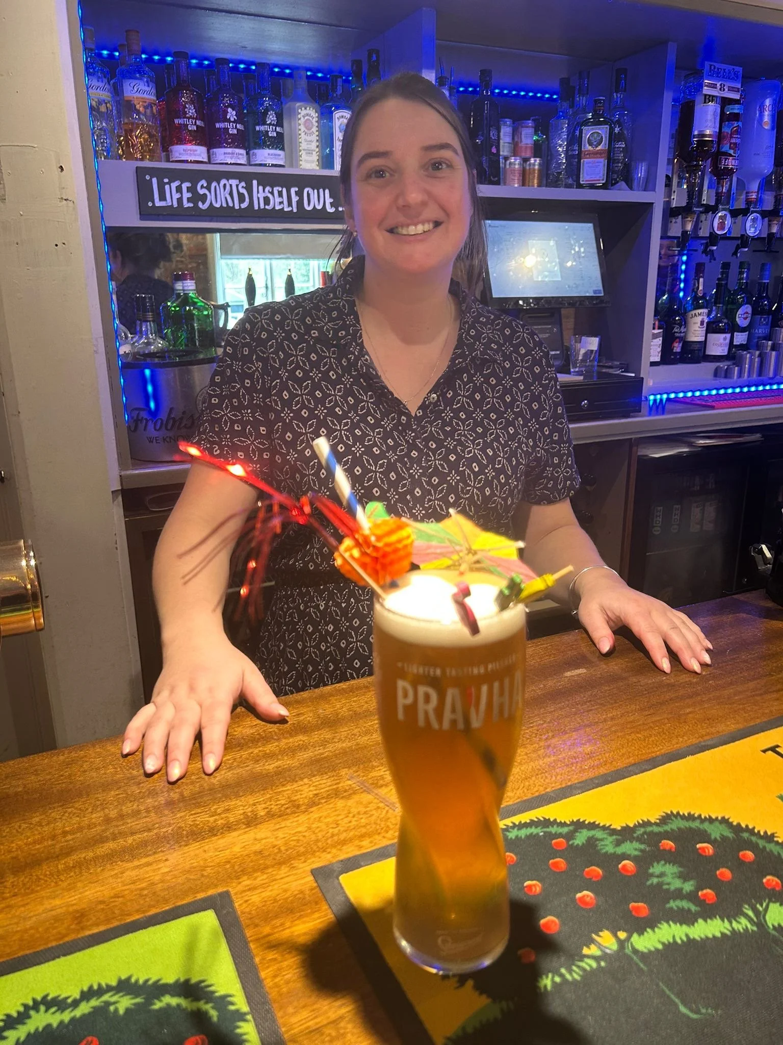 A woman behind a bar smiling, with a drink in the foreground decorated with a small umbrella and birthday candle. The bar has bottles of alcohol on shelves behind her, with a sign that says, 'Life sorts itself out.'