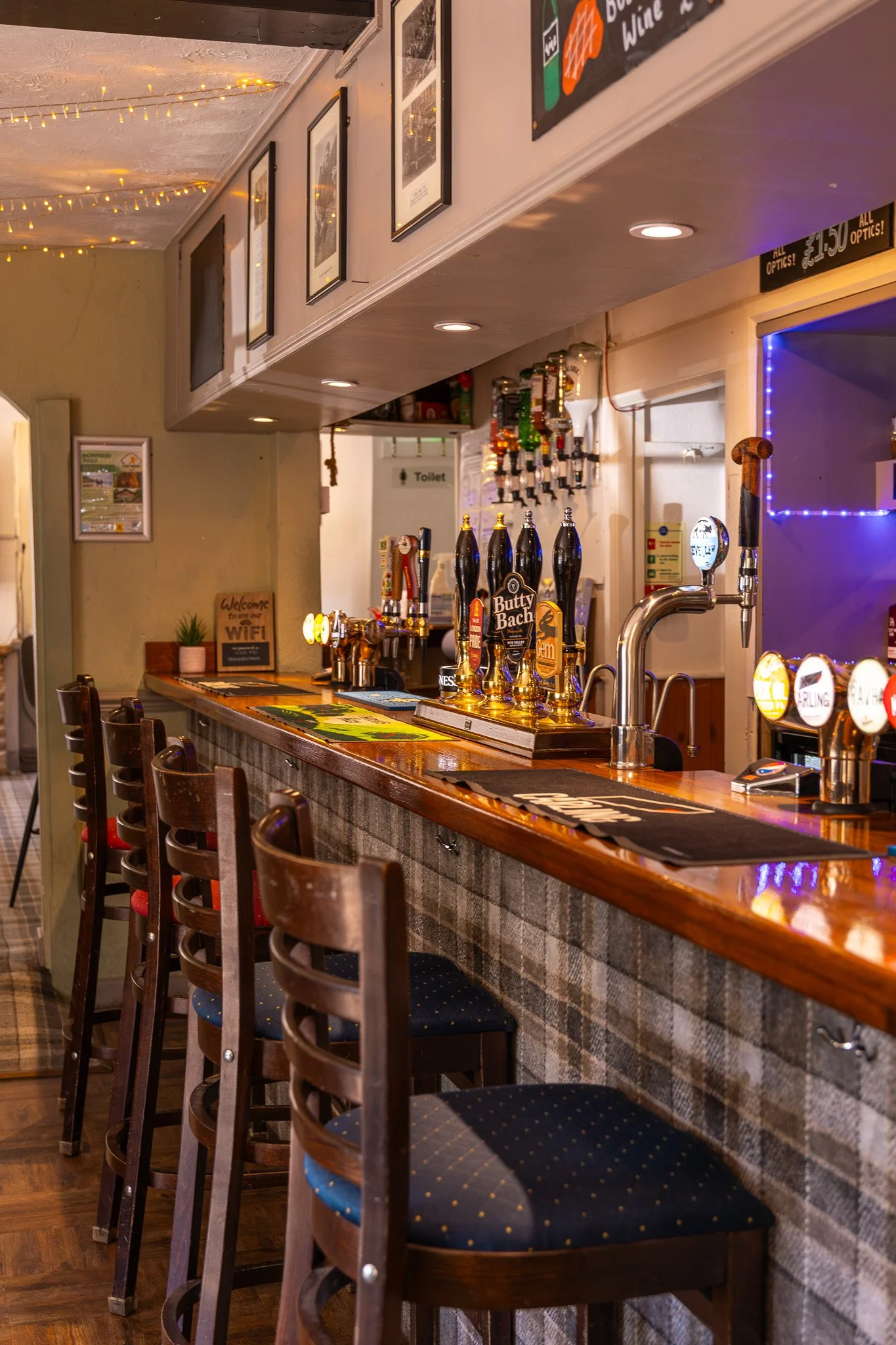 Interior of a bar with wooden chairs lined up along a bar counter, beer taps, and various decorations including framed pictures, fairy lights, and a chalkboard menu.