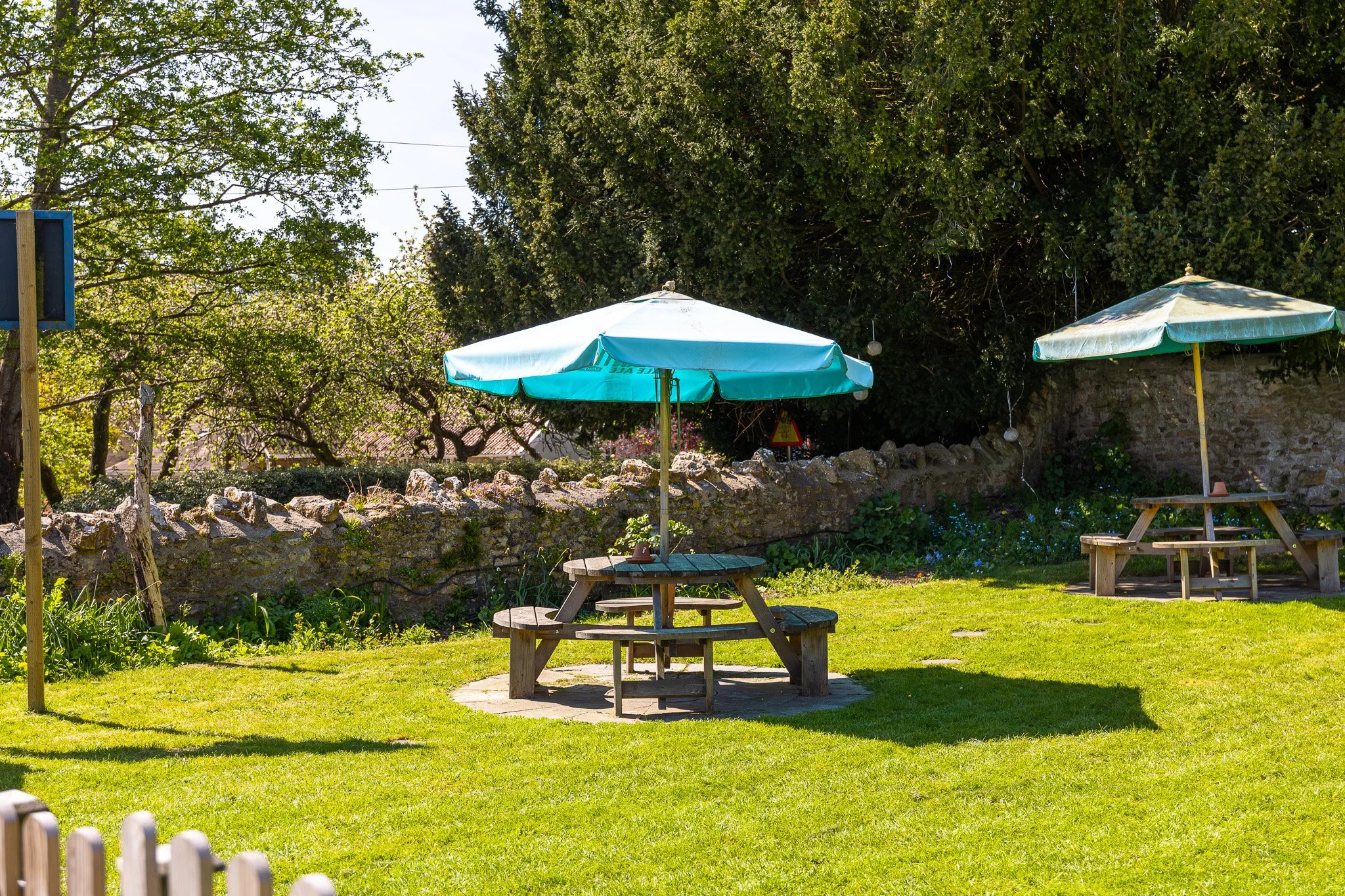 An outdoor garden with two wooden picnic tables and umbrellas, surrounded by trees, a stone wall, and lush grass.