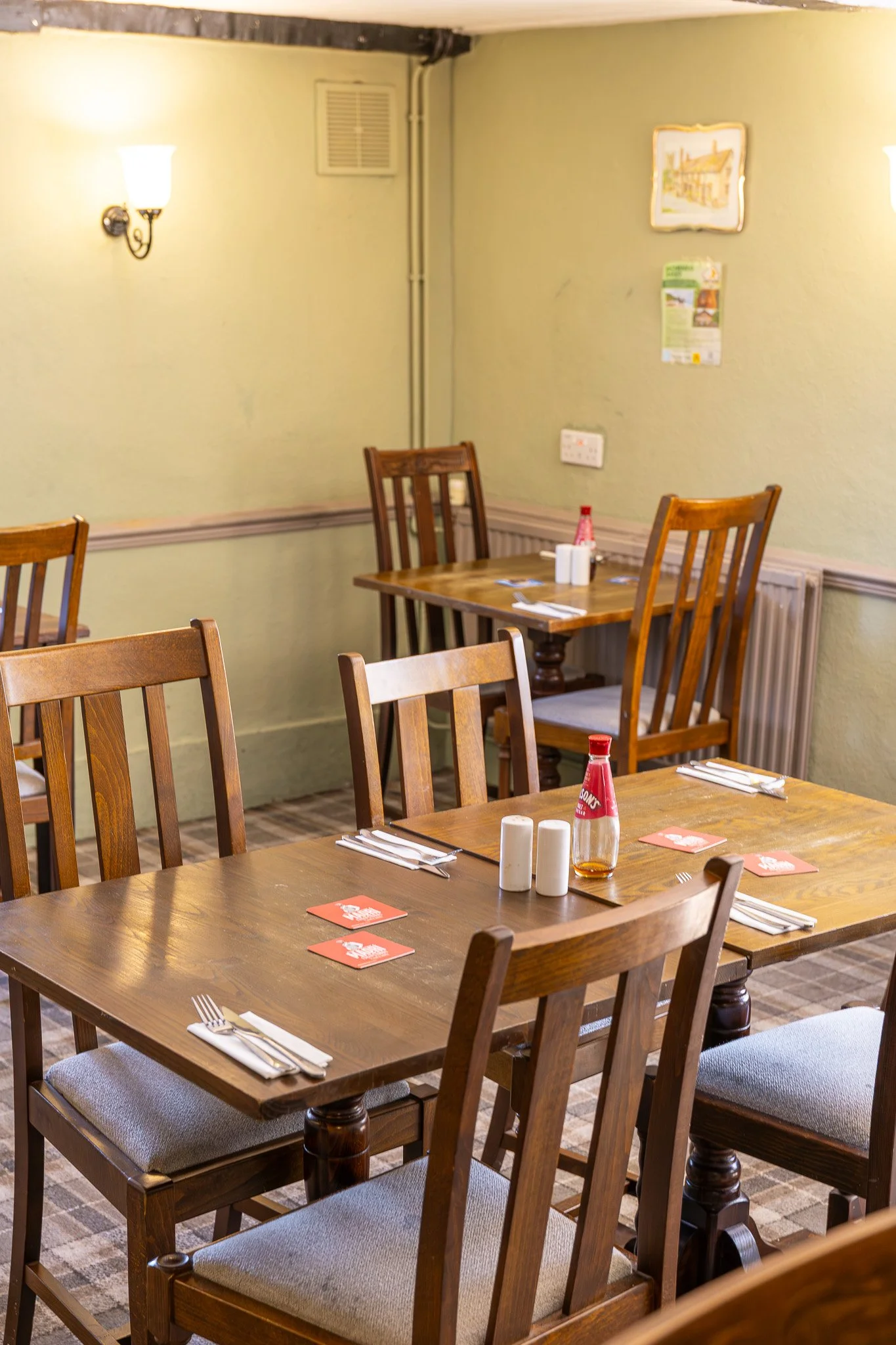 A cozy restaurant with wooden tables and chairs, set for dining with napkins, utensils, and condiments. The walls are painted light green with framed pictures and a lamp providing warm lighting.