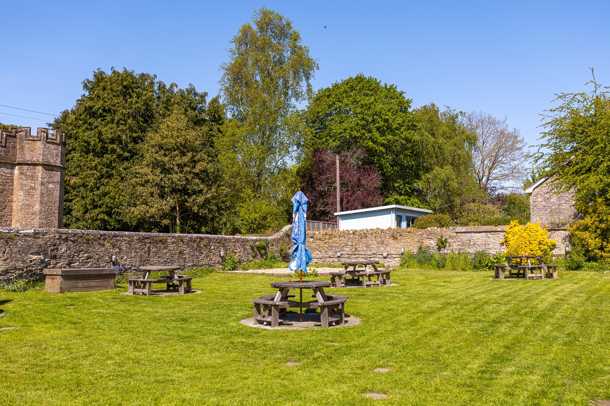 A sunny outdoor garden with wooden picnic tables and a closed blue and white striped umbrella, a stone wall, a grassy area, and green trees against a clear blue sky.