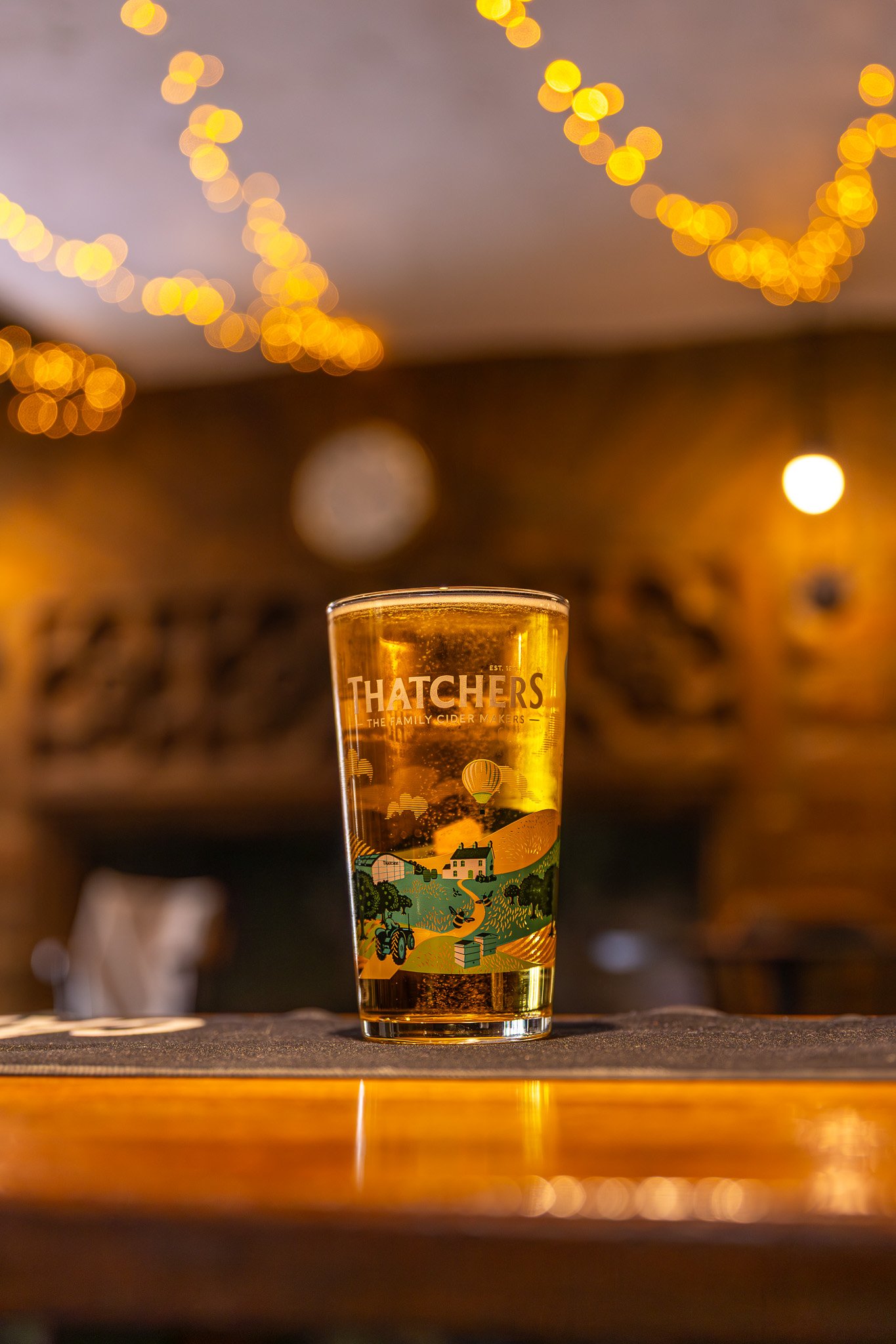 A pint glass of cider with a colorful illustrated label of a farm scene, set on a wooden surface with warm, bokeh light decorations in the background, creating a cozy ambiance.