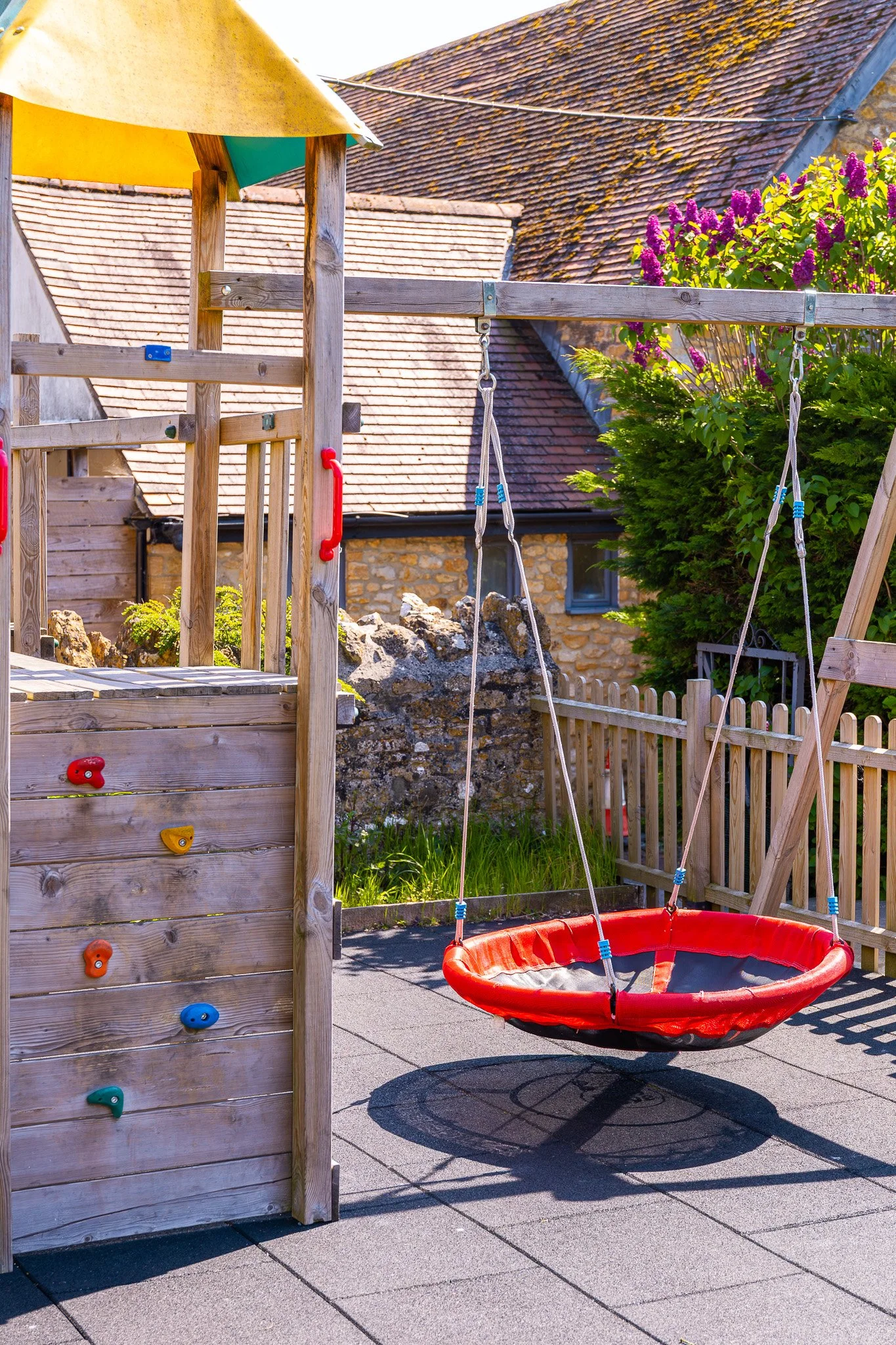 Child's outdoor play area with climbing wall, tire swing, and wooden fence, enclosed by brick and stone houses with tiled roofs.