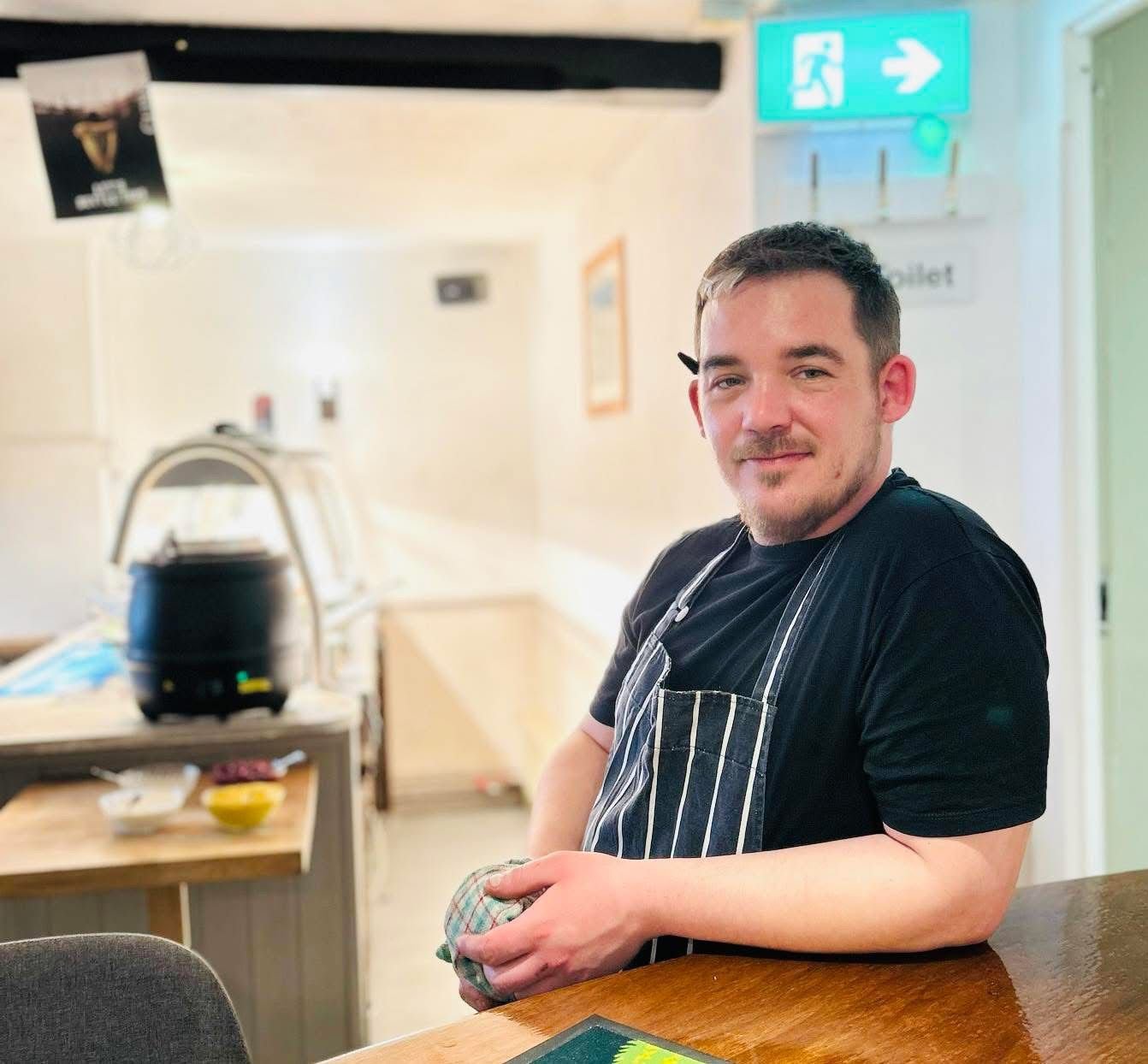 A man with short dark hair and a beard, wearing a black T-shirt and a striped apron, standing at a bar. He is holding a cloth and smiling at the camera. There is a carvery unit in the background