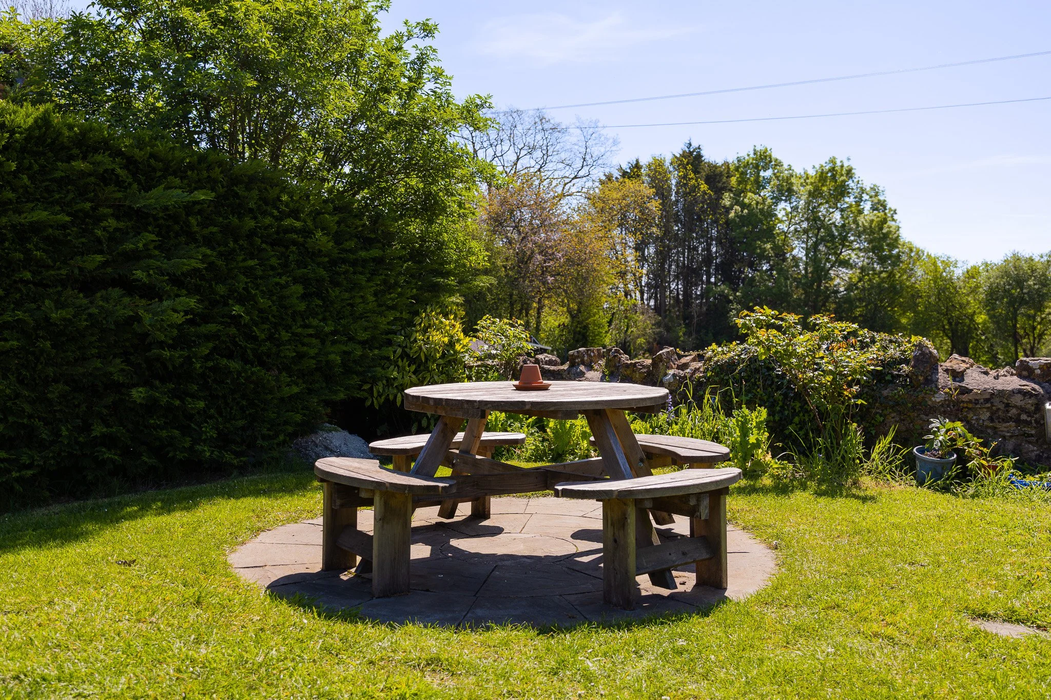 A wooden picnic table with attached benches in a grassy yard, surrounded by trees and bushes on a sunny day.