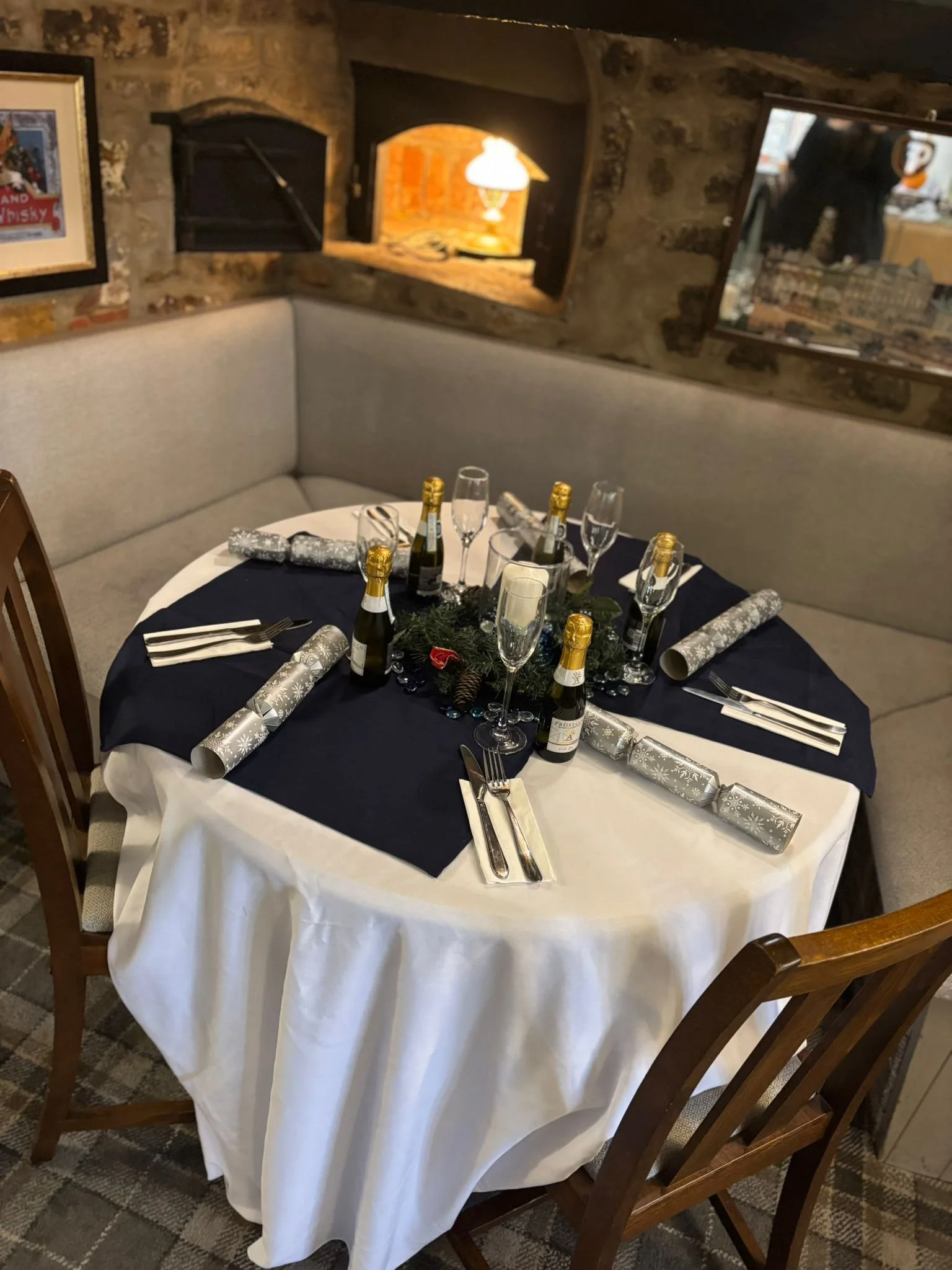A round dining table set for a celebration, with a dark blue tablecloth, white undercloth, champagne bottles, glasses, and silver-wrapped presents, in a rustic restaurant with exposed brick walls.