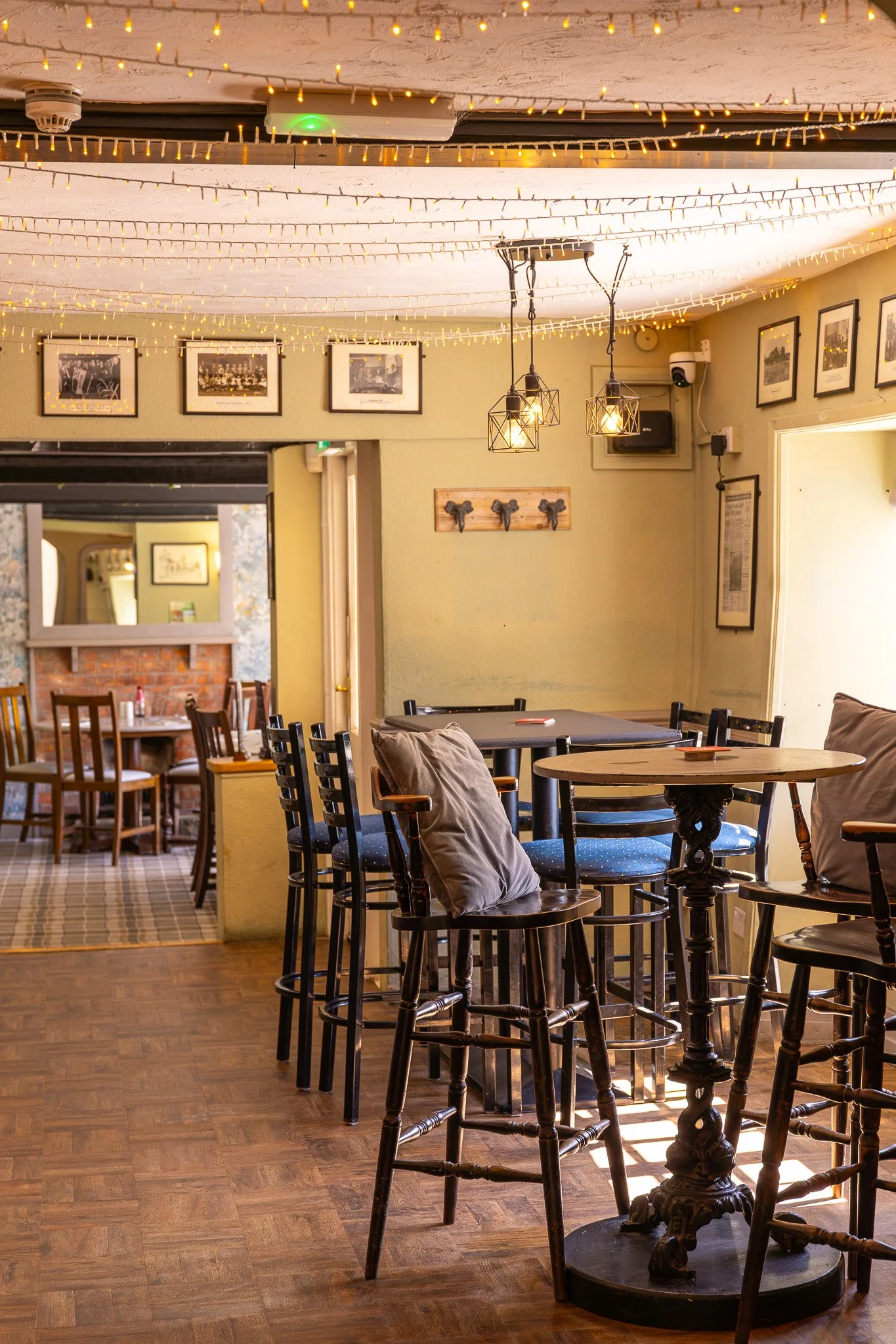 Interior of a cozy restaurant with wooden tables and chairs, fairy lights strung across the ceiling, framed black-and-white photos on the walls, and decorative pendant lights hanging from the ceiling.