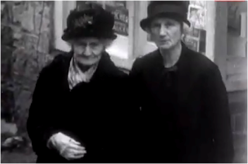 Two women standing close together outdoors, both wearing dark clothing and hats, in black-and-white photograph.