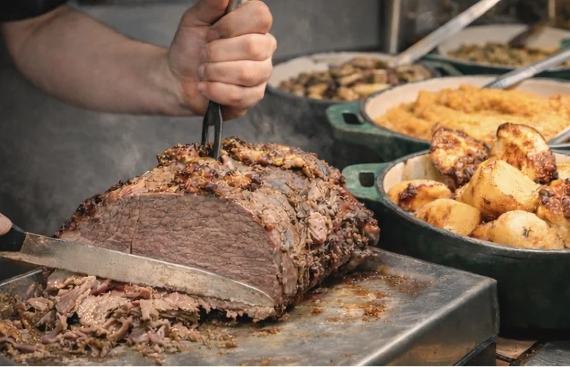 Person carving a large cooked beef roast with sides of potatoes and vegetables in the background.