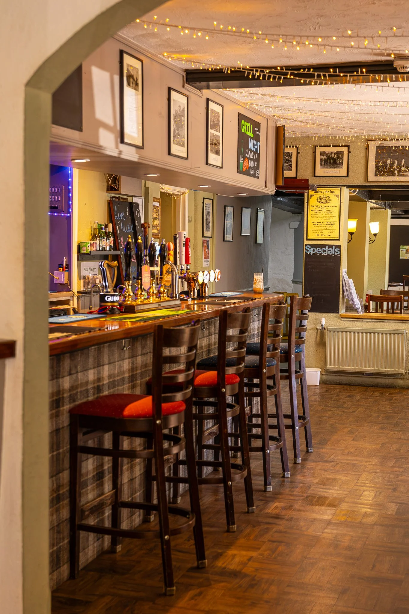 Interior of a bar or pub with a wooden bar counter, high chairs, beer taps, and various framed pictures on the wall. String lights hang from the ceiling, creating a warm atmosphere.