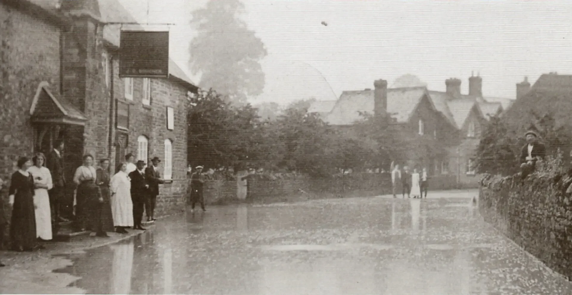 A black-and-white photo of a street scene with people standing along a curb and walking in the street, in front of houses with chimneys, trees, and a cloudy sky.