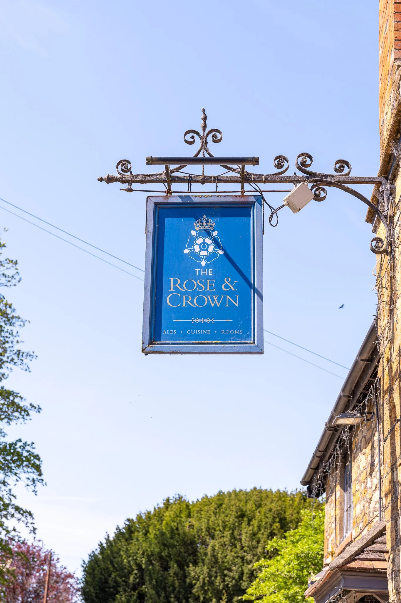 Blue hanging sign with a rose and crown logo and the text 'The Rose & Crown', advertising ales, cuisine, and rooms, attached to a metal bracket on the side of a building with a clear blue sky in the background.