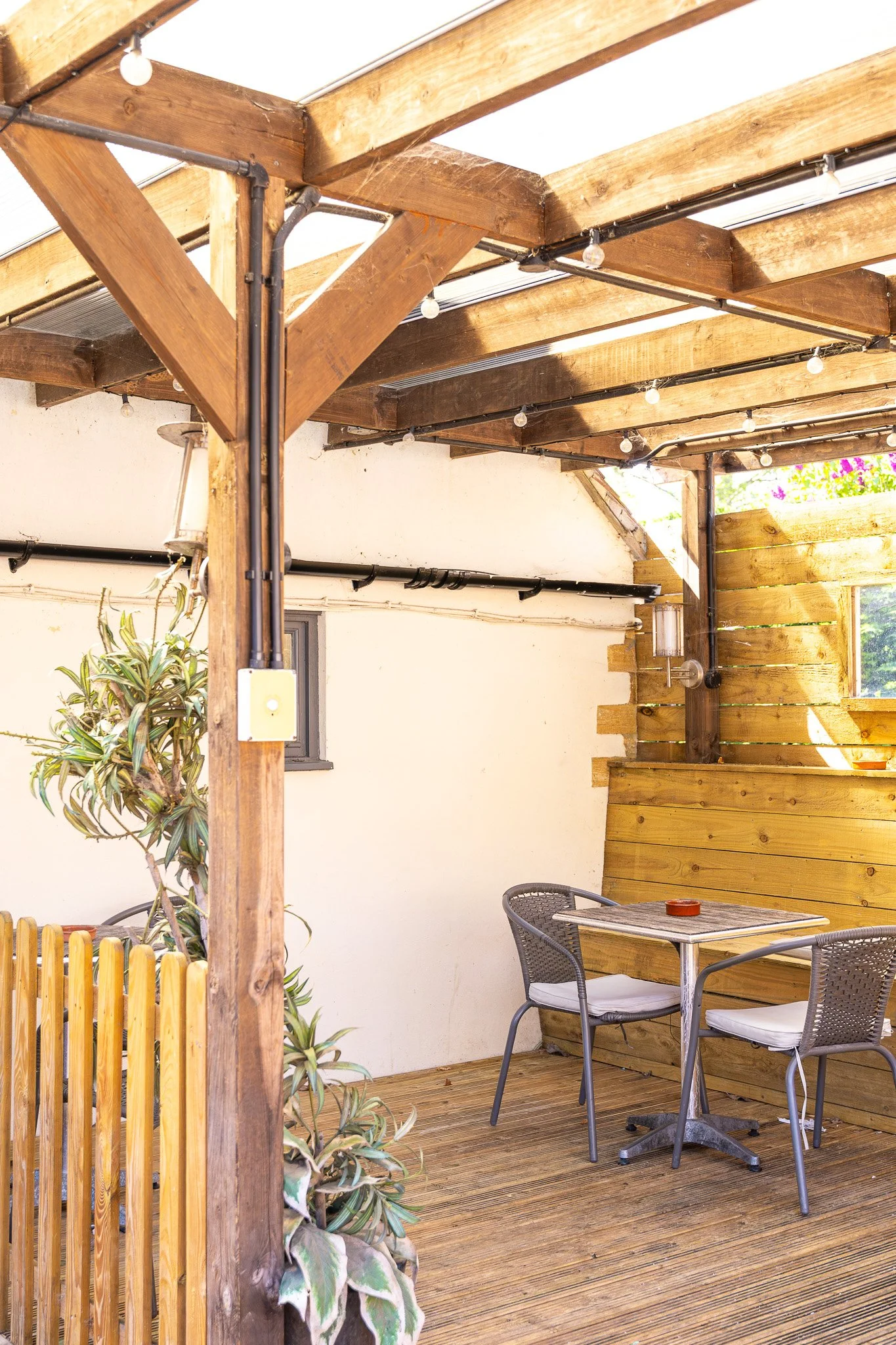 A shaded outdoor patio area with a wooden pergola, string lights, small round tables, two black chairs with cushions, and a potted plant.