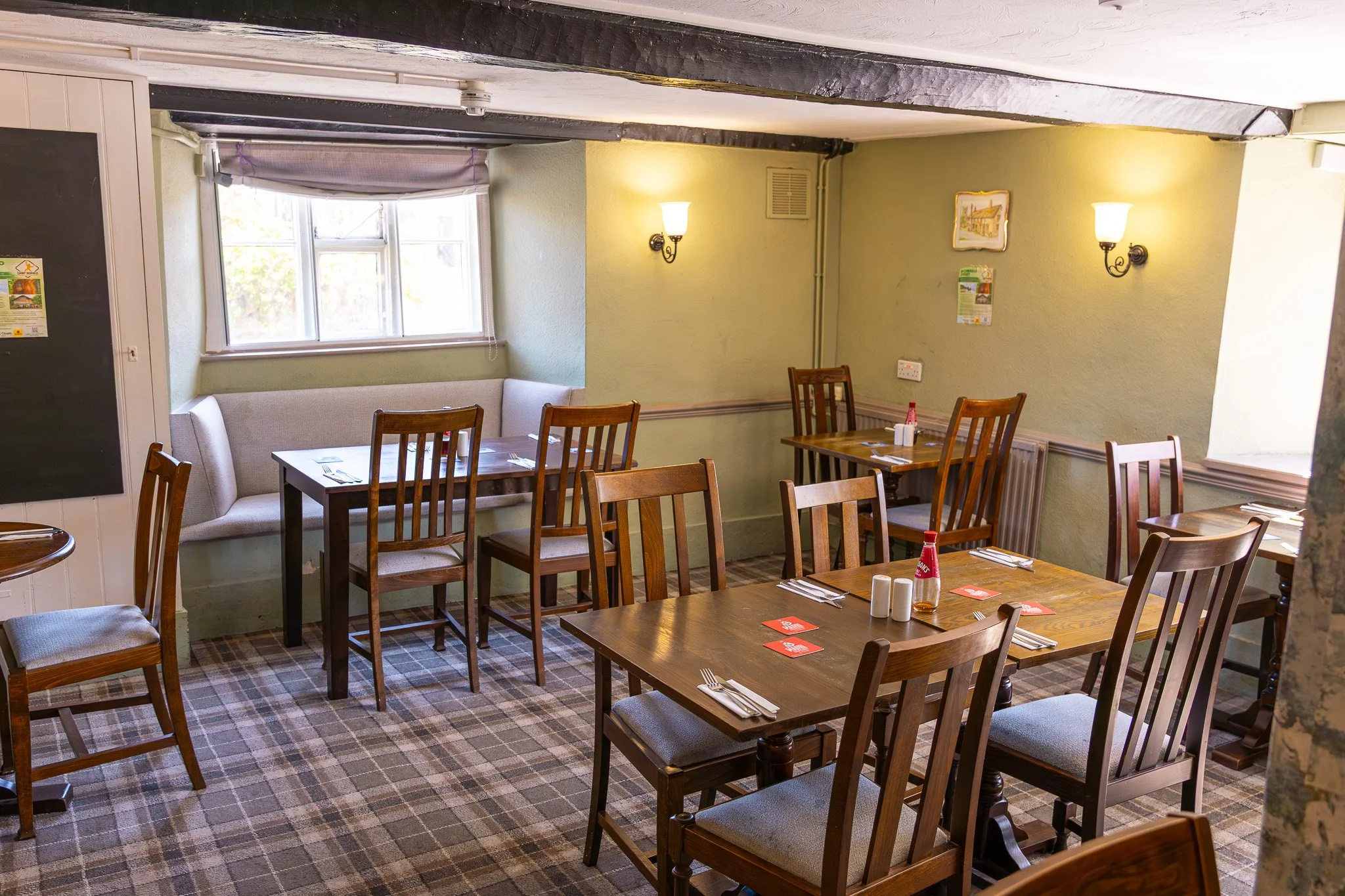 Interior of a cozy restaurant with wooden tables and chairs, green walls, wall-mounted lamps, a window, and a plaid carpet.