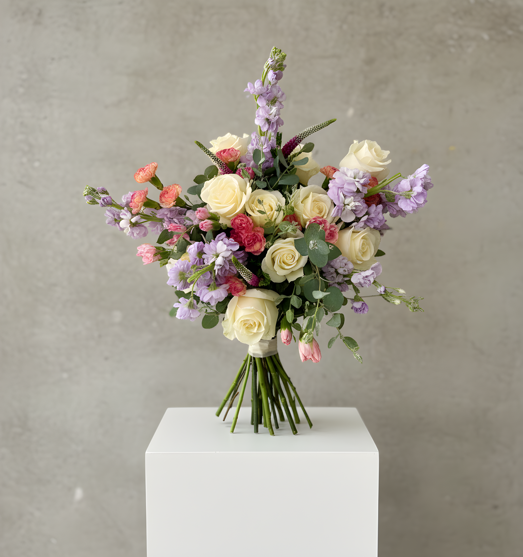 A mixed bouquet of white roses, purple snapdragons, pink screamers, and greenery arranged in a white vase on a white table against a gray background.