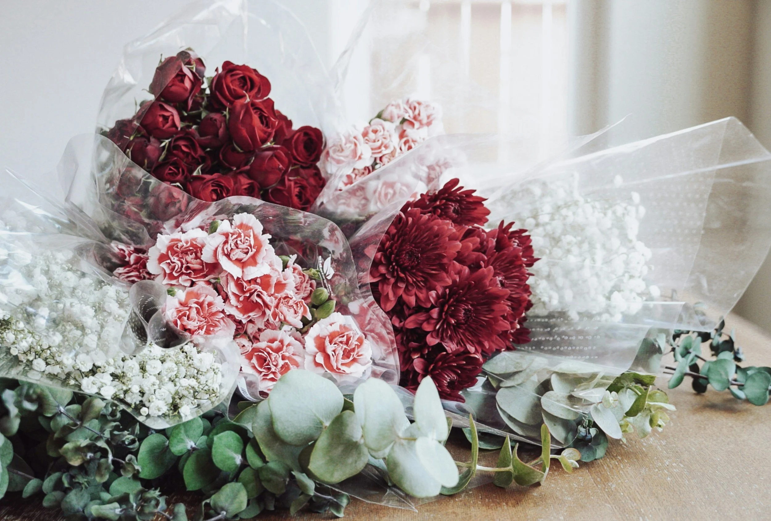 Bouquets of red, pink, and white flowers, including roses and carnations, with green foliage, wrapped in clear plastic, placed on a wooden surface.