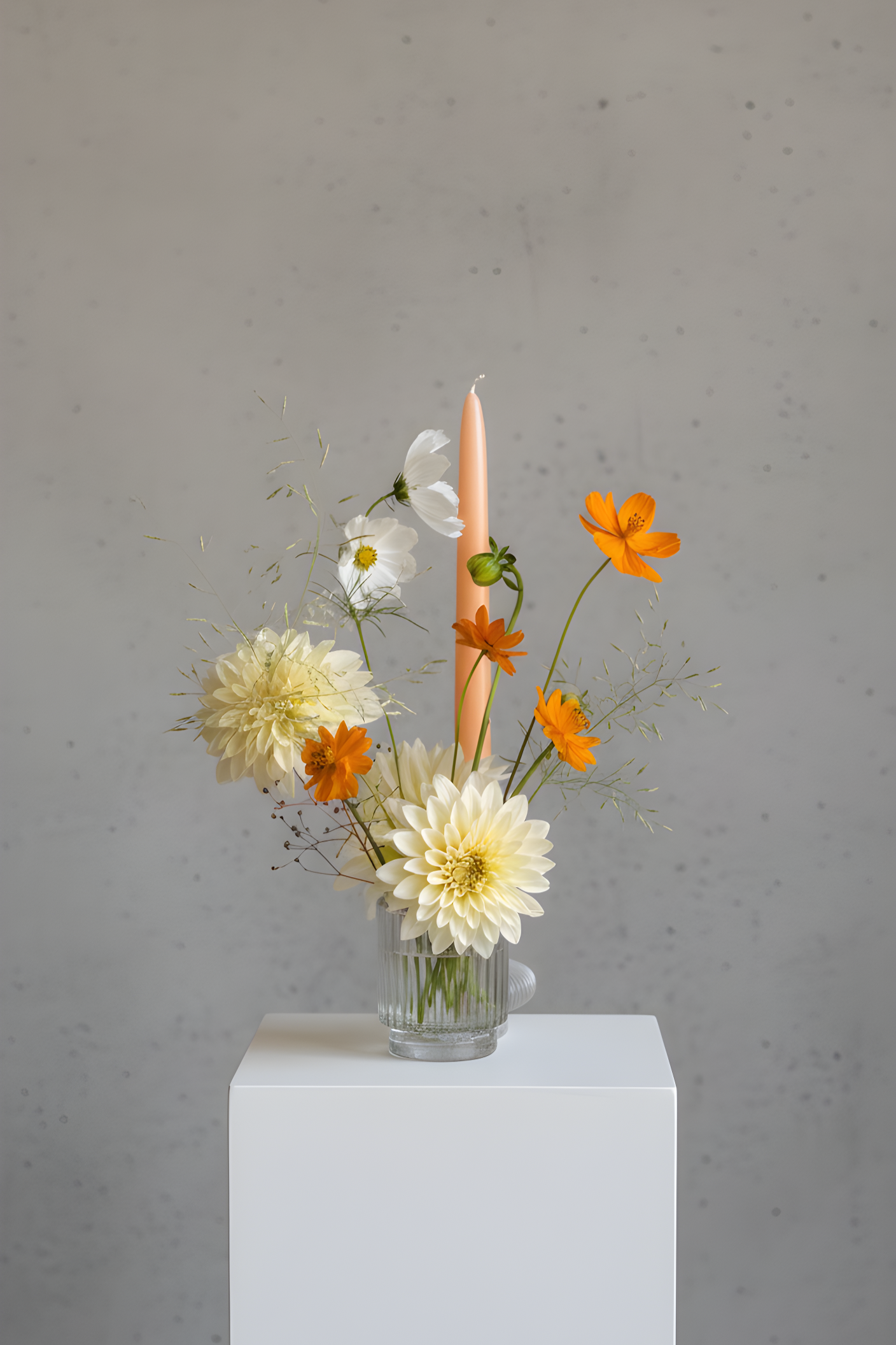 A floral arrangement in a clear glass vase with white, orange, and cream flowers, a peach-colored candle, and green foliage, placed on a white pedestal against a gray textured background.