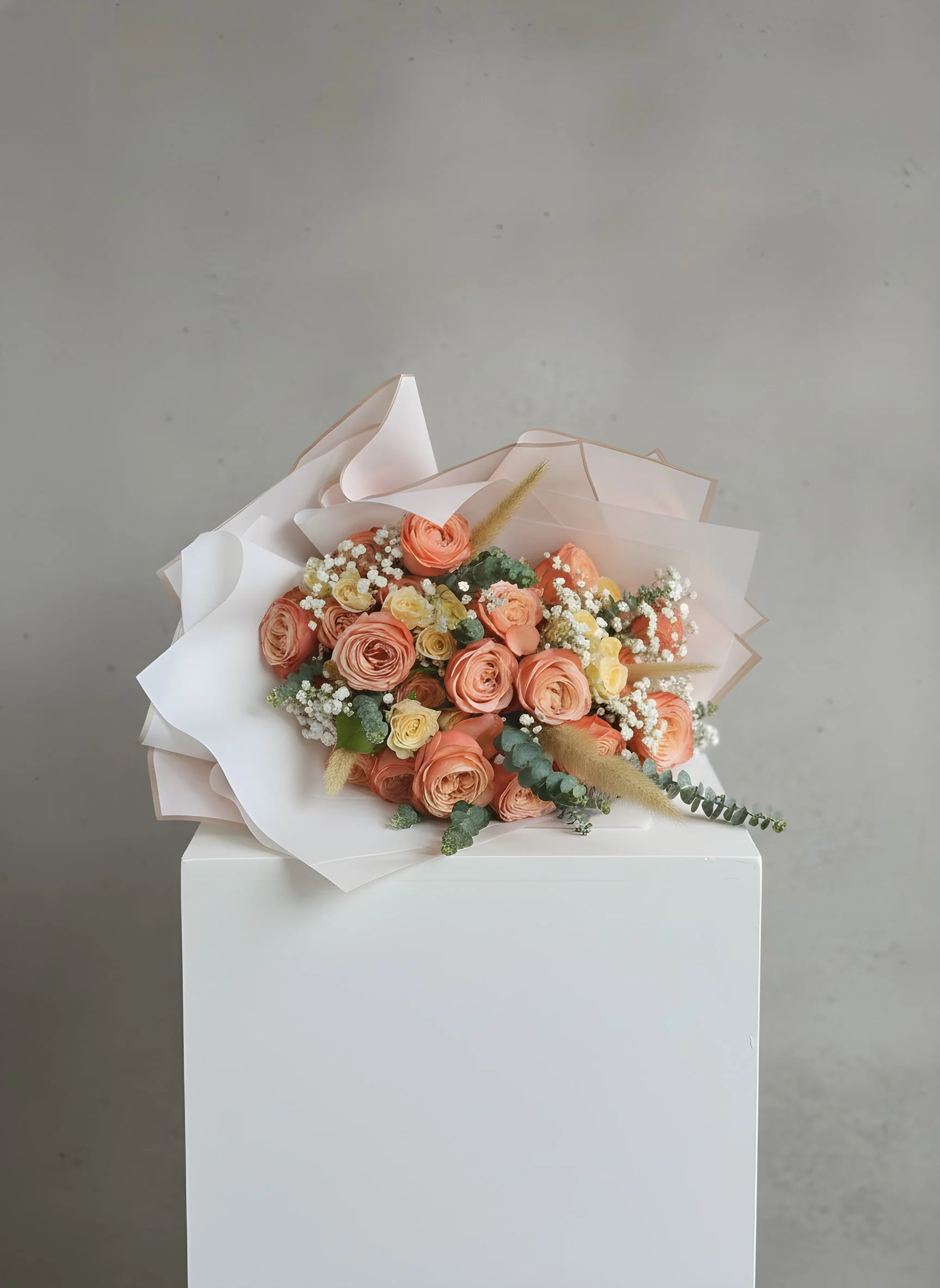 A bouquet of peach, yellow, and white roses with greenery and small white filler flowers wrapped in white and translucent paper, placed on a white pedestal against a gray background.