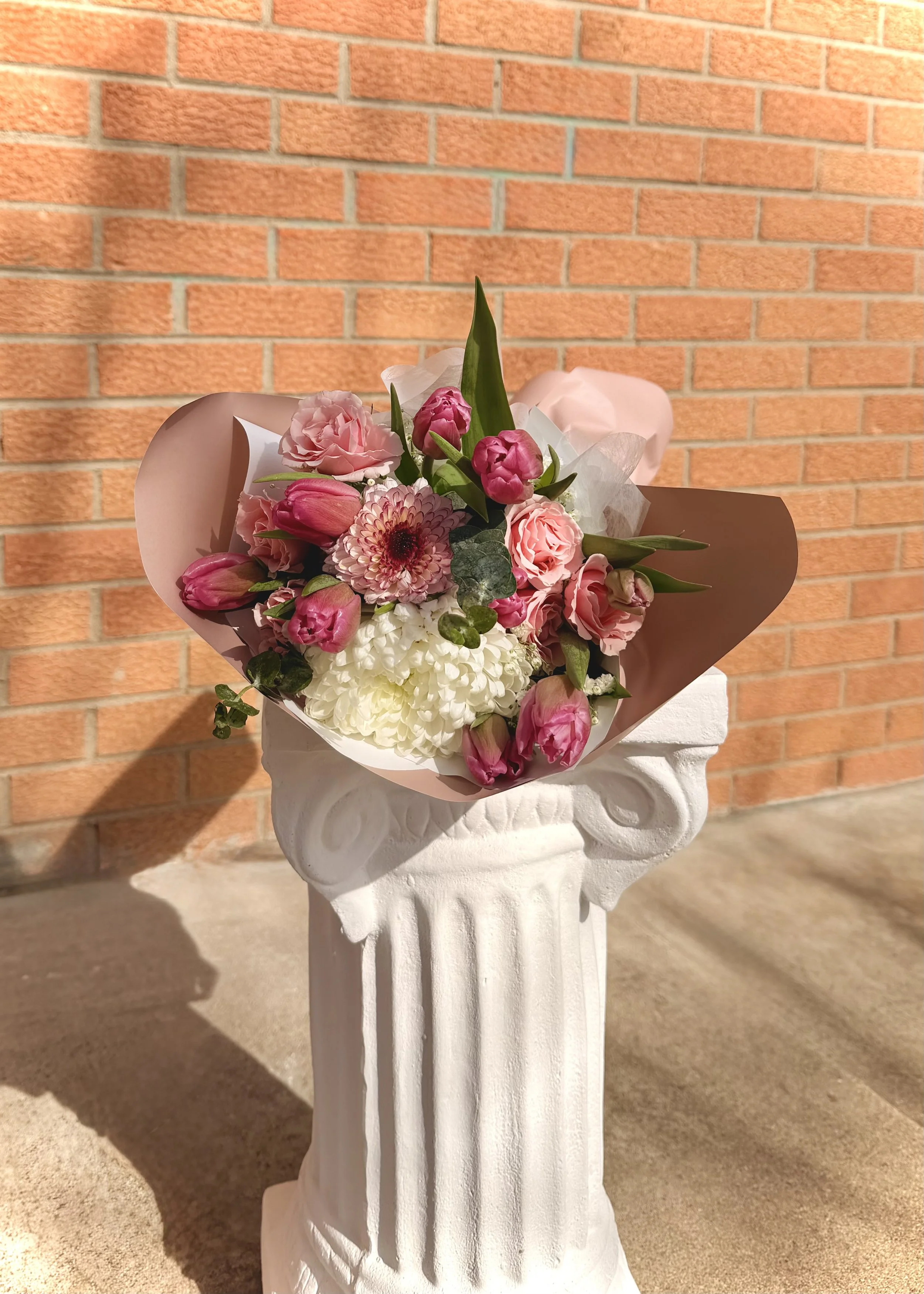 A bouquet of pink and white flowers, including tulips and roses, arranged with green leaves, wrapped in pink paper, placed on a white decorative pedestal against a brick wall background.