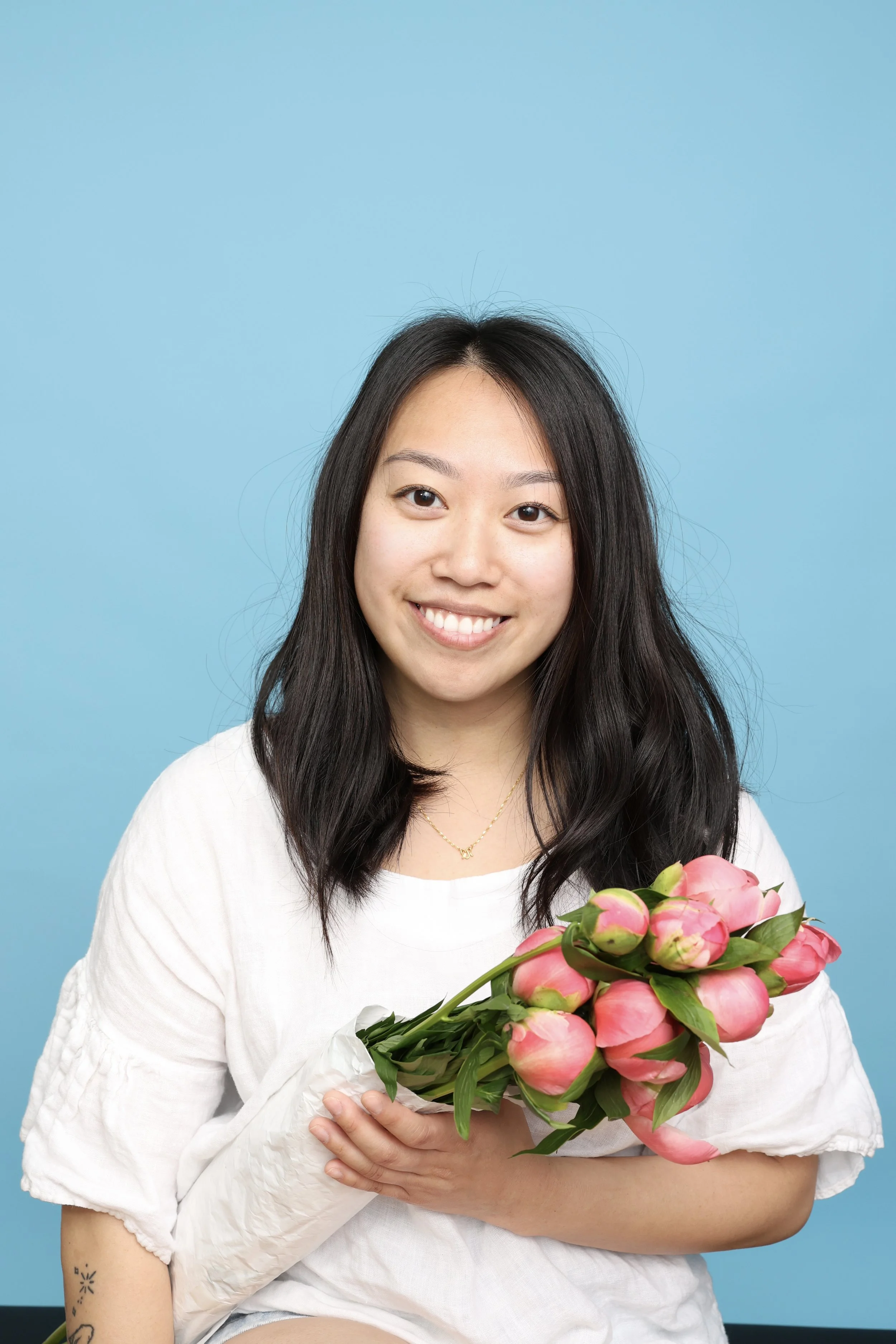 A young woman with long black hair, smiling, holding a bouquet of pink peonies against a blue background.