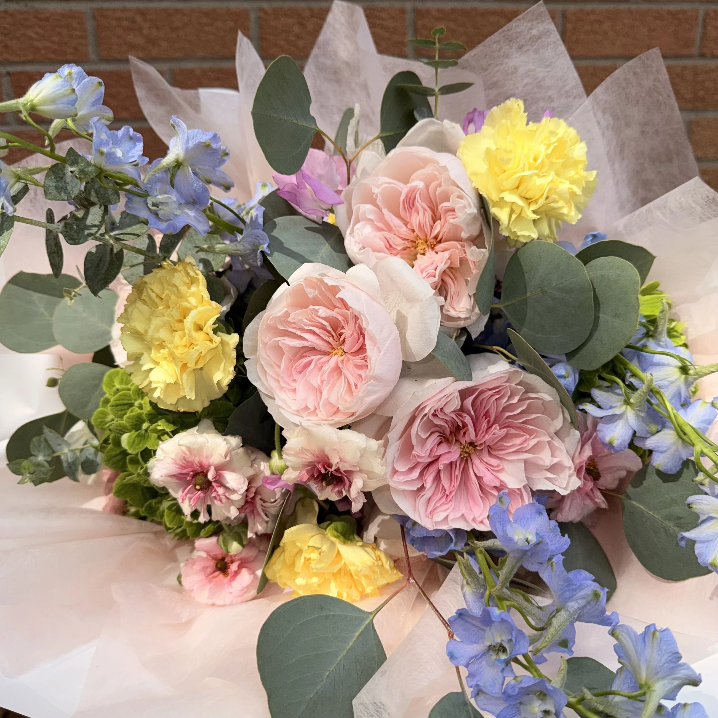 A bouquet of pastel-colored flowers including pink, yellow, and purple blooms with green leaves, wrapped in white tissue paper, set against a brick wall background.