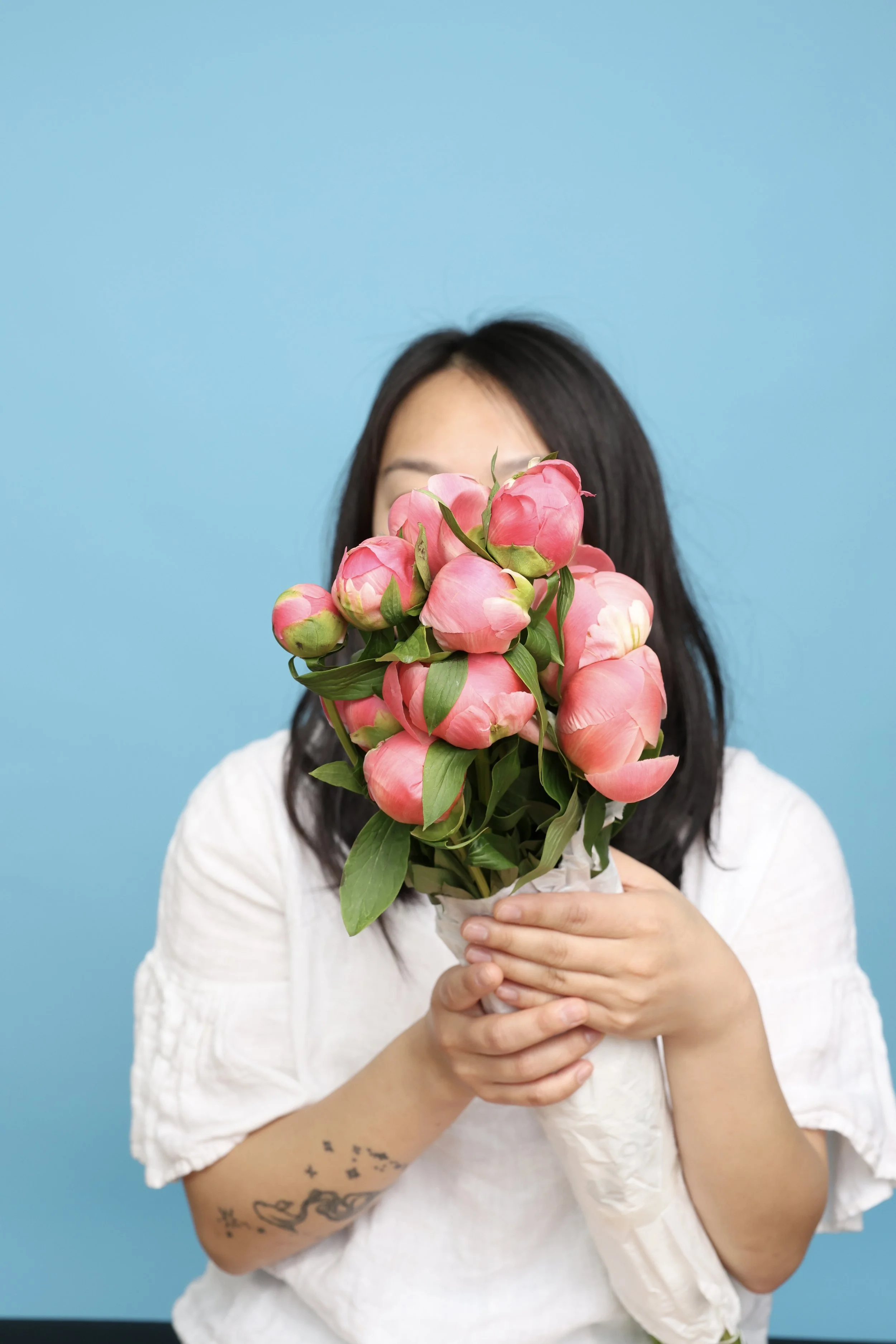 A woman with dark hair in a white shirt holds a bouquet of pink peonies in front of her face, against a light blue background.