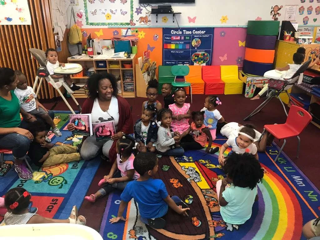 A teacher reading a book to a group of children in a colorful classroom, children sitting on a vibrant alphabet carpet, some sitting on chairs, others on the floor, engaging with the story.