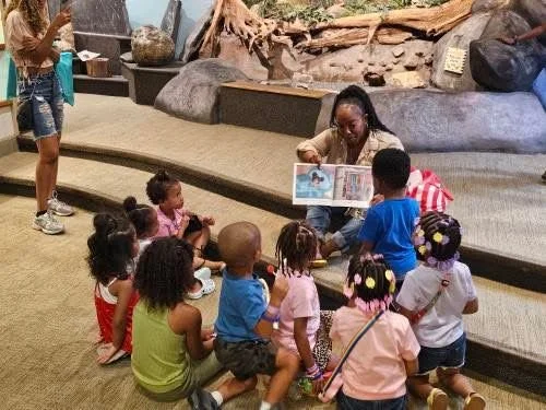 A woman reading a book aloud to a group of young children sitting on the floor in an indoor setting. The children are attentively listening, and a second woman stands nearby.