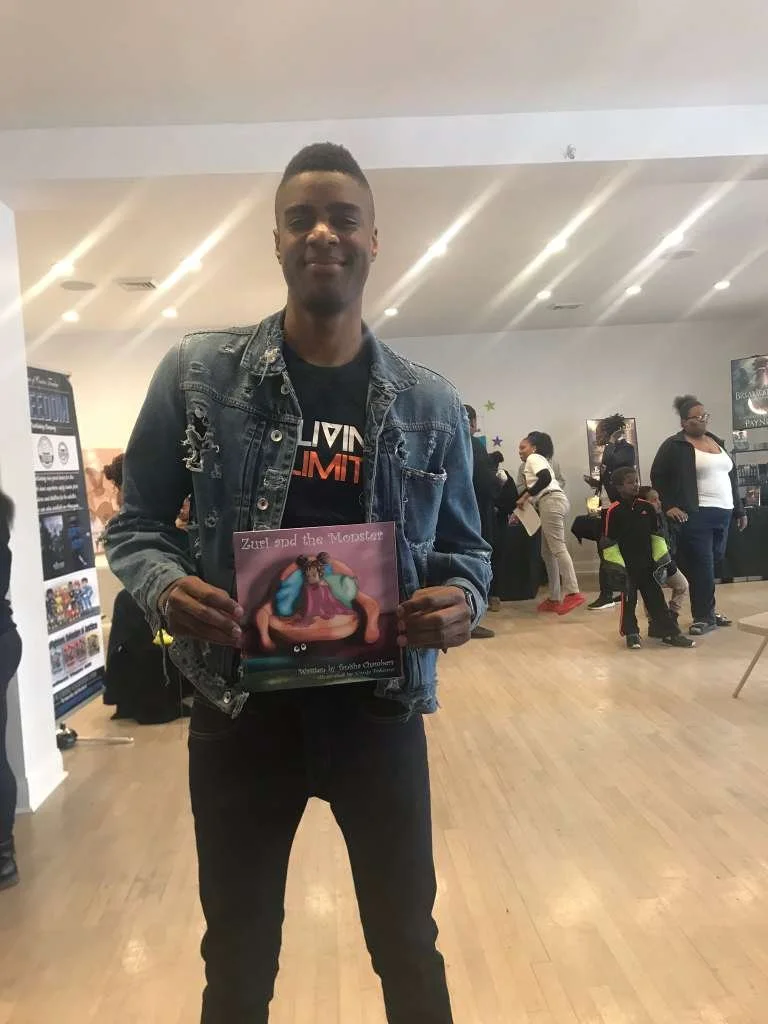 Young man wearing a denim jacket and black T-shirt, holding a children's book titled 'Zuzu and the Monster,' standing in a busy indoor space with multiple people and displays in the background.