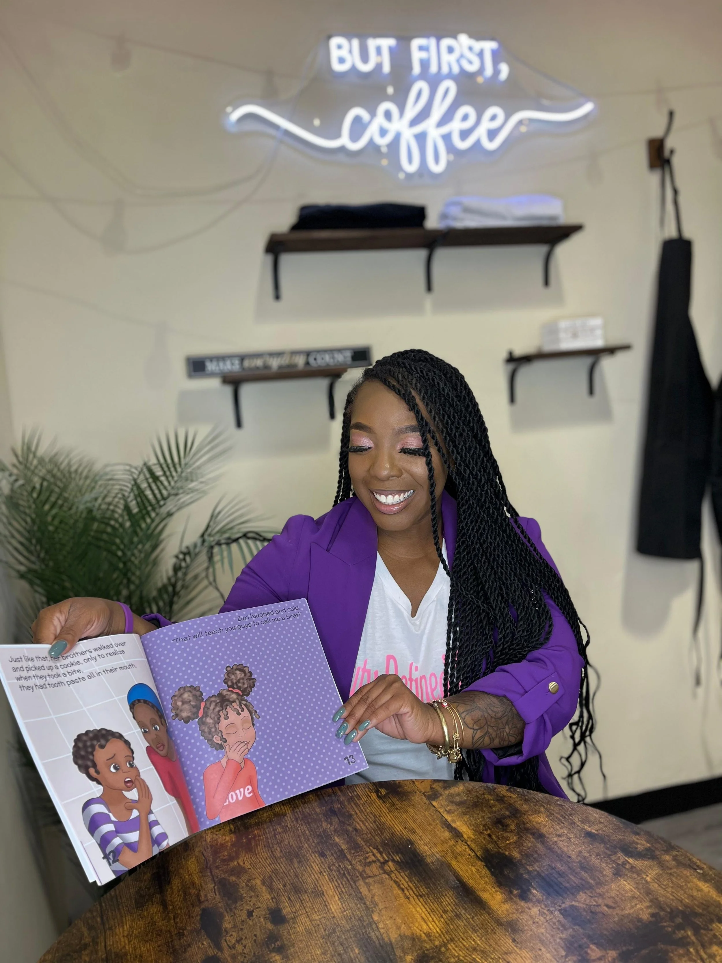 A smiling woman with braided hair wearing a purple blazer and white t-shirt looking at a children's book with colorful illustrations of children, sitting at a wooden table in a cafe or bookstore. There is a neon sign reading 'But First, Coffee' on th