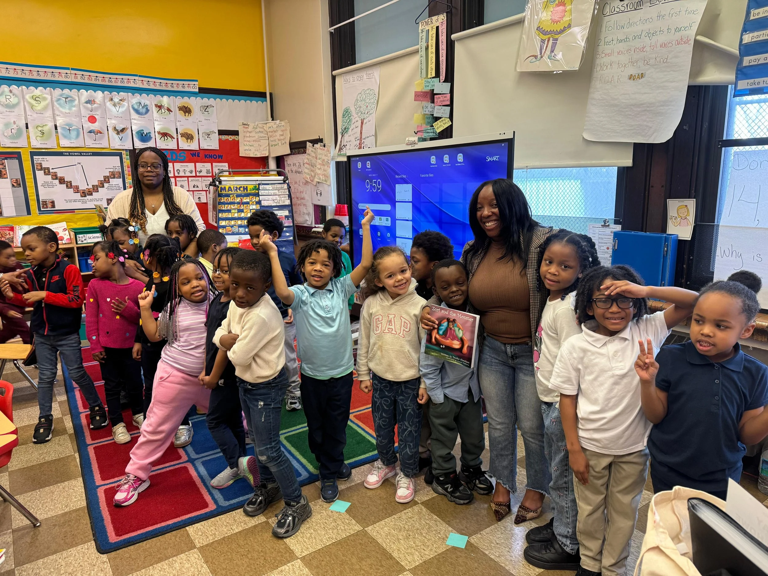 A group of young children standing in a classroom, smiling and posing for the photo, with two women, likely teachers, standing behind them. The classroom has colorful posters and educational materials on the walls, including a rainbow rug on the floo