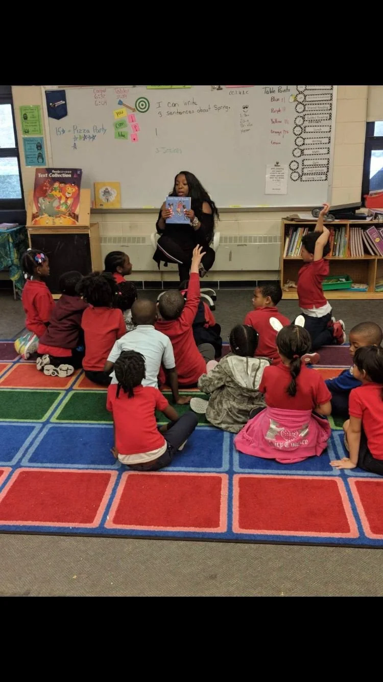 A classroom with a teacher reading a book to children sitting on a colorful carpet. Several children are raising their hands as they listen.