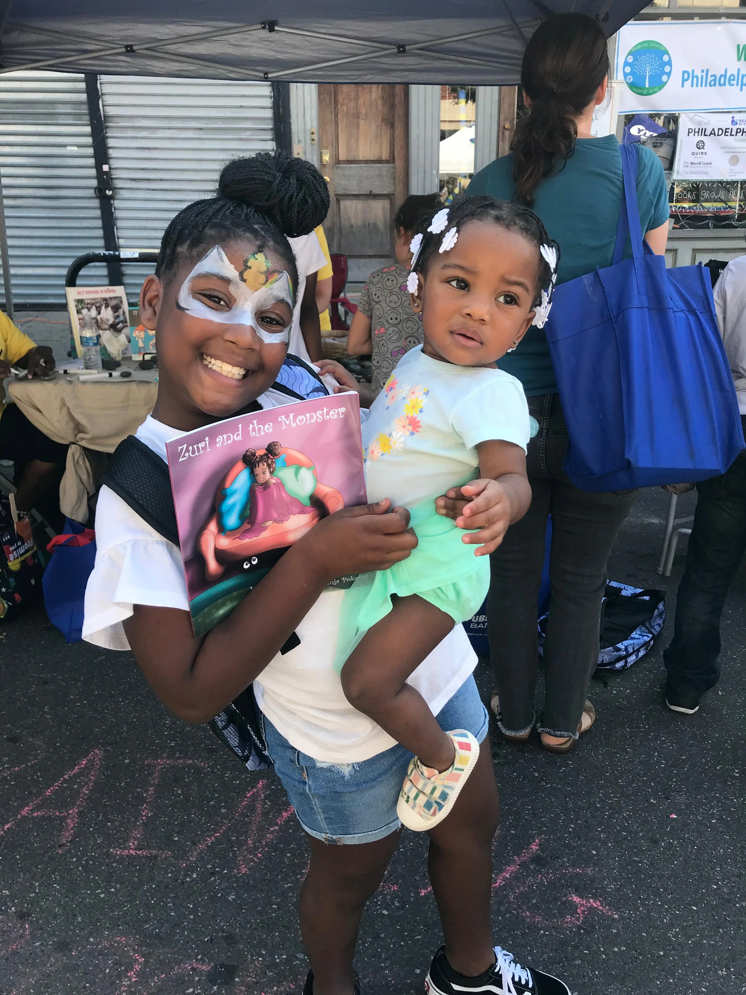 Two young girls at an outdoor event, one with face paint and the other holding a children's book titled 'Zuri and the Monster'. The girl with face paint is smiling, wearing a white shirt, denim shorts, and black sneakers. The girl holding the book ha