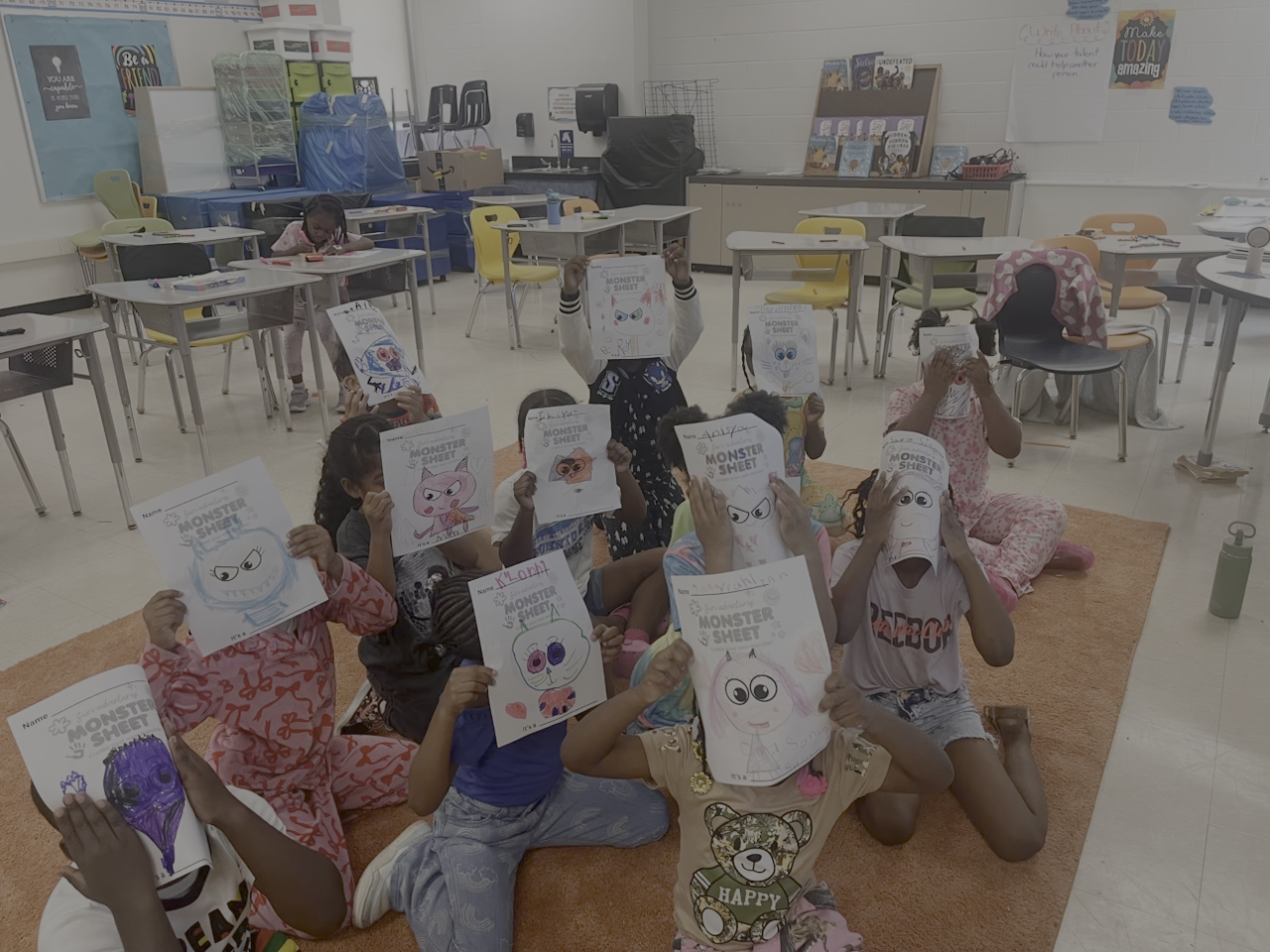 Children sitting on a carpet in a classroom, holding up drawings of monsters they have colored and decorated.