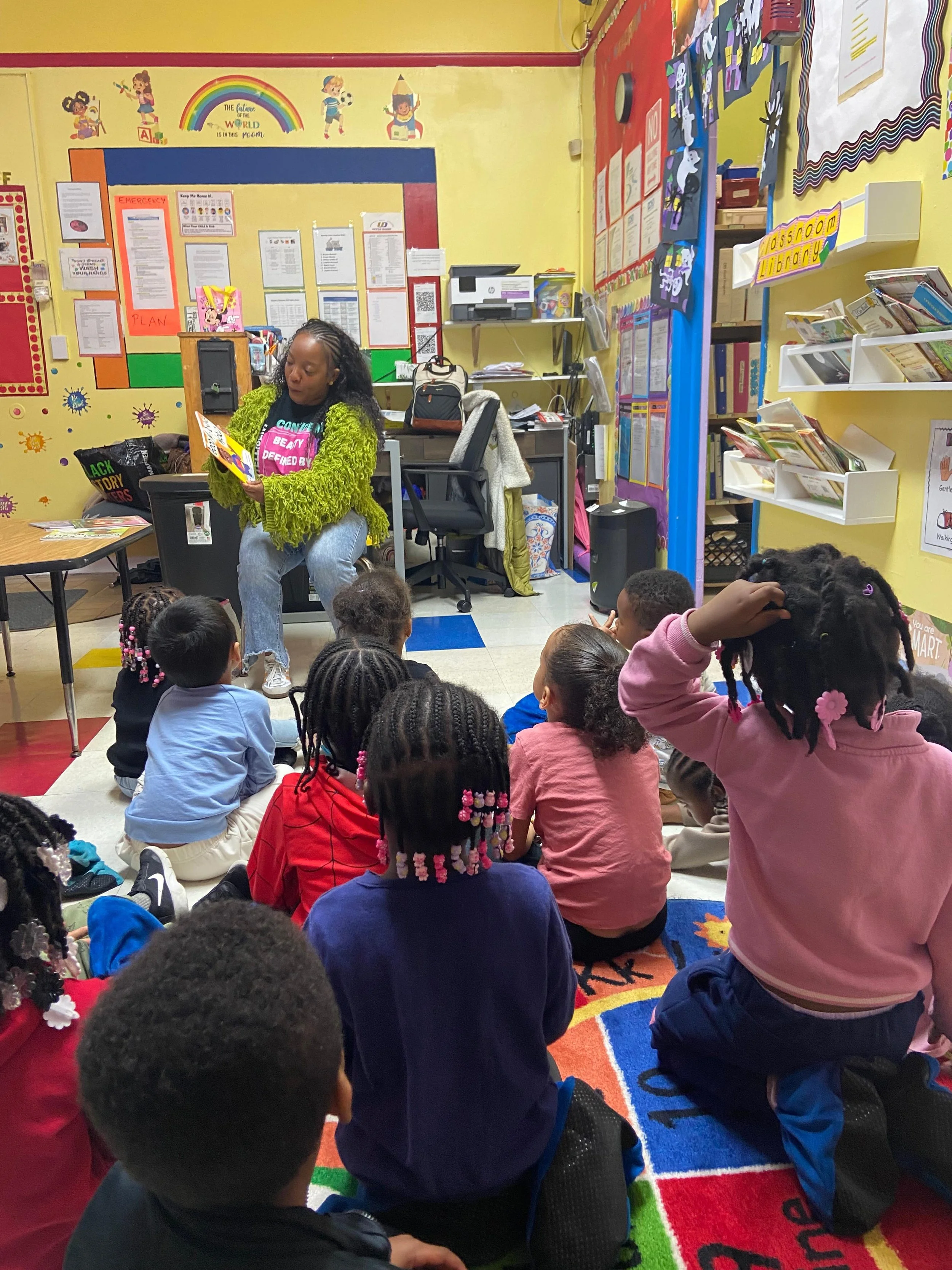 A woman reading a book to a group of children sitting on a colorful classroom rug. The classroom has yellow walls decorated with children’s artwork, educational posters, and a rainbow mural.