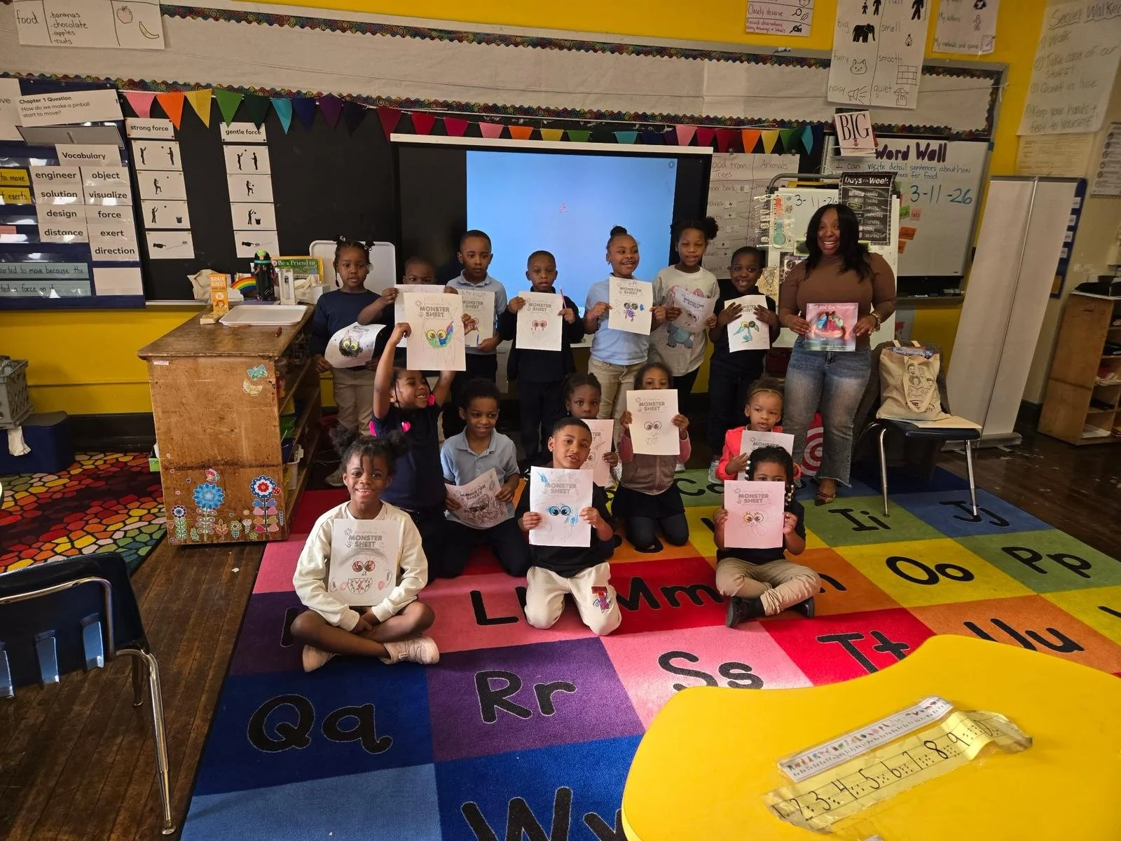 A classroom with children holding up kids' drawings of monsters with big eyes and teeth, sitting and standing on a colorful alphabet rug, with a teacher smiling on the right side.