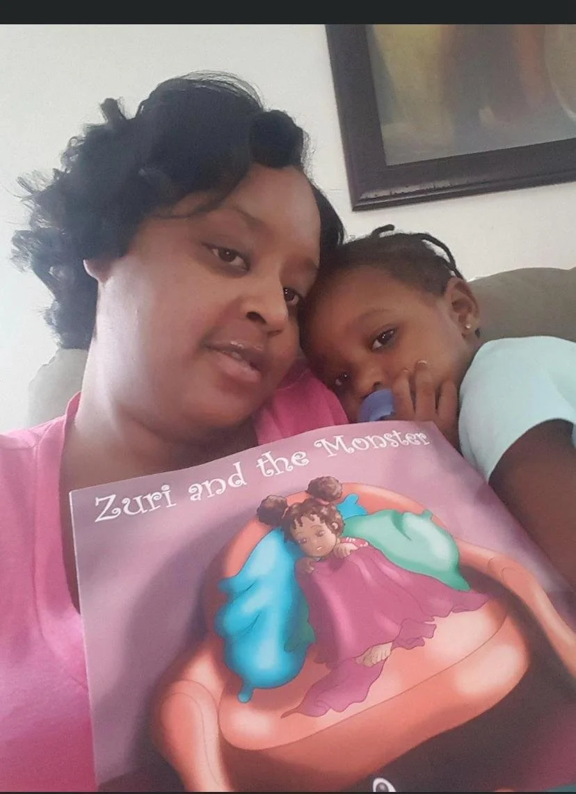 A woman with short, curly black hair holding a children's book titled "Zuri and the Monster," sitting with a young girl who is resting her head on the woman's shoulder, both looking at the camera.