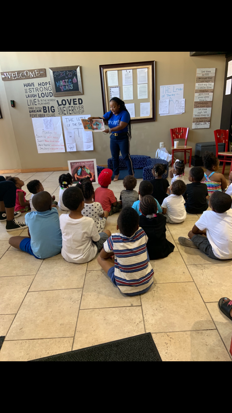 A woman is reading a children's book aloud to a group of young children sitting on the floor in a room with beige walls, decorated with inspirational posters and written notes.