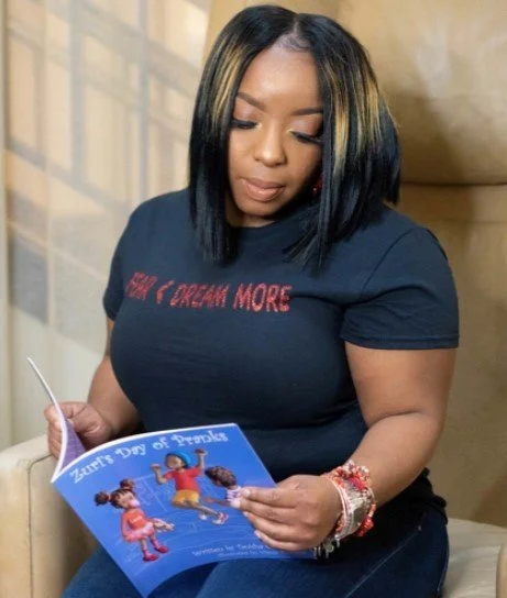 A woman with shoulder-length black hair with blonde highlights, wearing a navy T-shirt that says 'never stop dreaming more,' sitting and looking at a colorful children's book titled 'Turning the Dog of Dreams.'