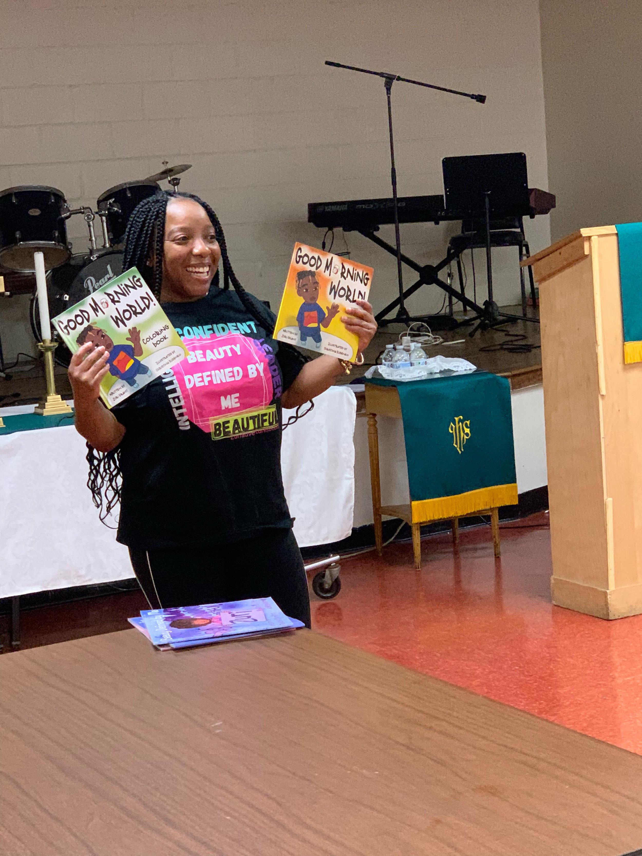 A woman smiling and holding two children's books titled "Good Morning World!" in a room with musical instruments, a podium, and some pamphlets resting on a table.