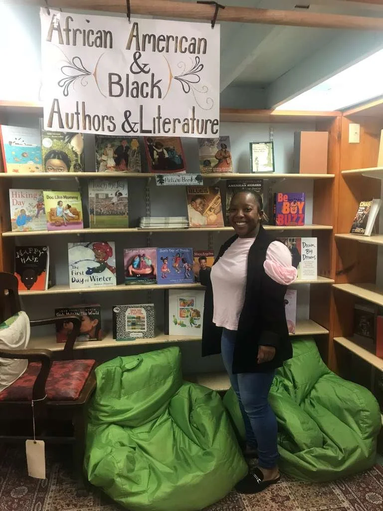 A woman standing in front of a bookshelf labeled 'African American & Black Authors & Literature' in a library. She is smiling and wearing a white shirt with pink puffed sleeves, a black jacket, and jeans. There are green bean bag chairs and a wooden 