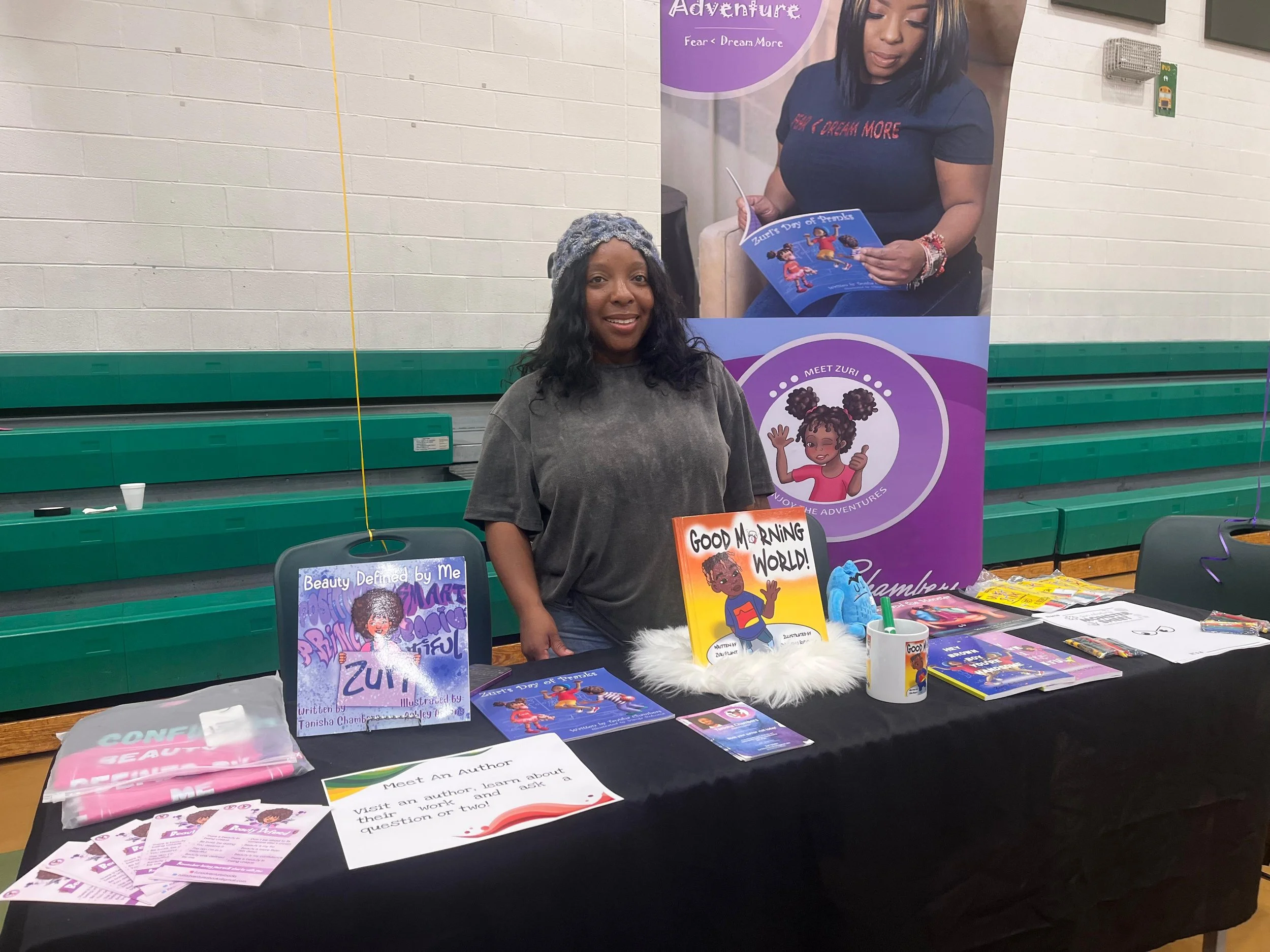 A woman with dark hair wearing a gray shirt standing behind a table at a book fair. The table displays children's books, pamphlets, and promotional materials about Zuri and her adventures. A large purple banner behind her features an illustration of 