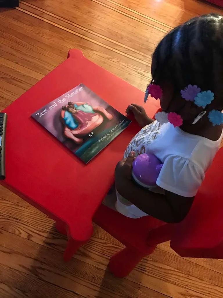 A young girl with colorful flower hair clips looks at a book titled "Zuri and the Monster" on a red table.