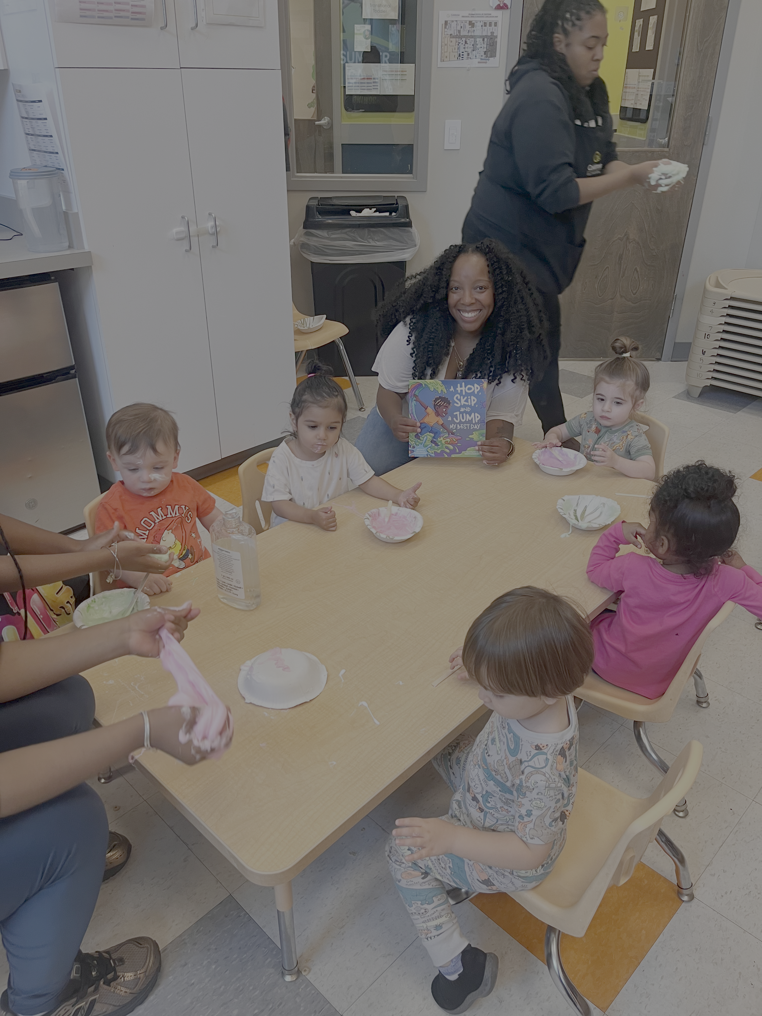 A group of young children sitting at a table with pink and white Slimes, with an Author Tanisha Chambers smiling and holding a children's book, in a daycare setting.