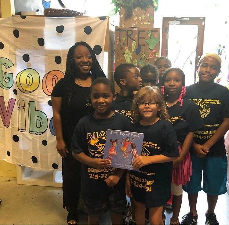 Group of children and an adult woman at a celebration, with a banner in the background that says 'Good Vibes'. One girl holds a book titled 'Zuri's Day of Dance'.