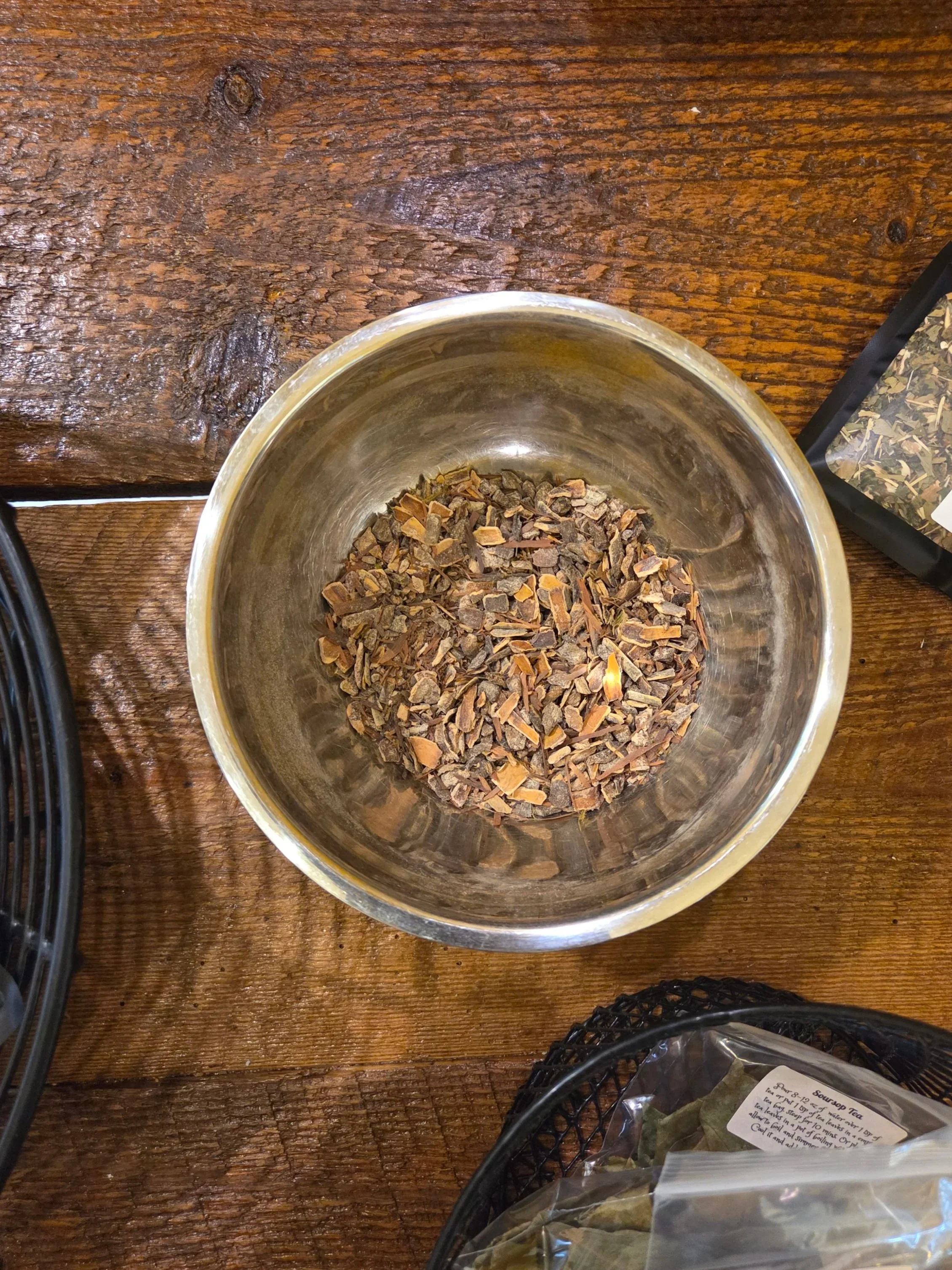 A metal bowl with dried, chopped herbs or bark placed on a dark wooden table. Herbal remedies and plant based medicines are crafted in-store. Using handpicked ingredients.
