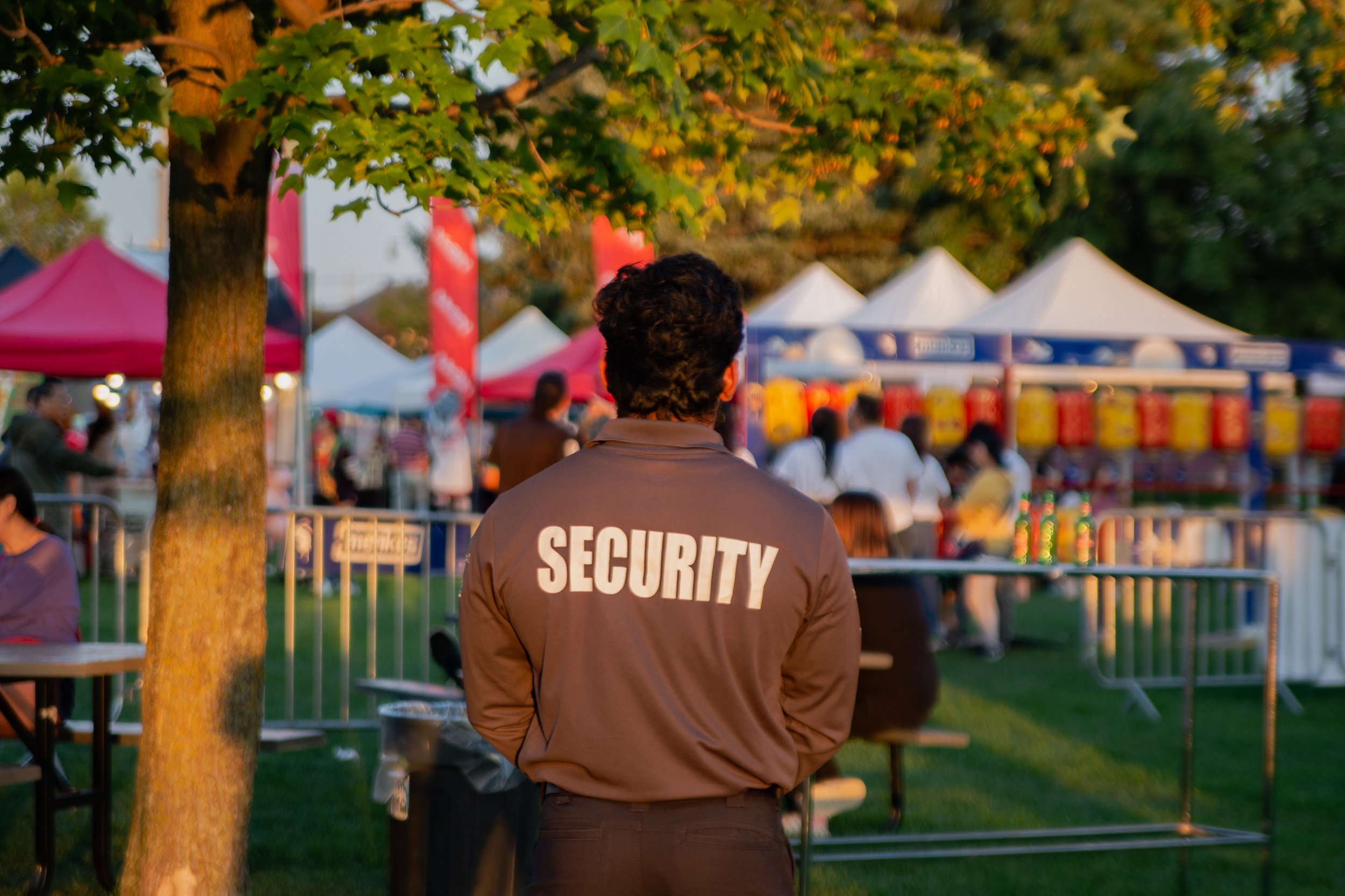 Security guard standing outdoors at a festival or fair, facing away from the camera, wearing a brown jacket with 'SECURITY' printed on the back, surrounded by tents and people.