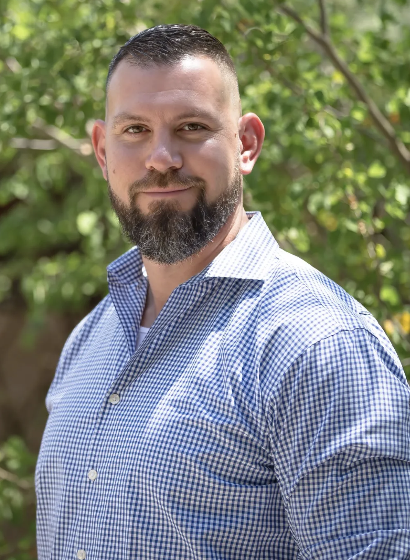 A man with a beard and short, dark hair wearing a blue and white checkered shirt, standing outdoors with a background of green trees.