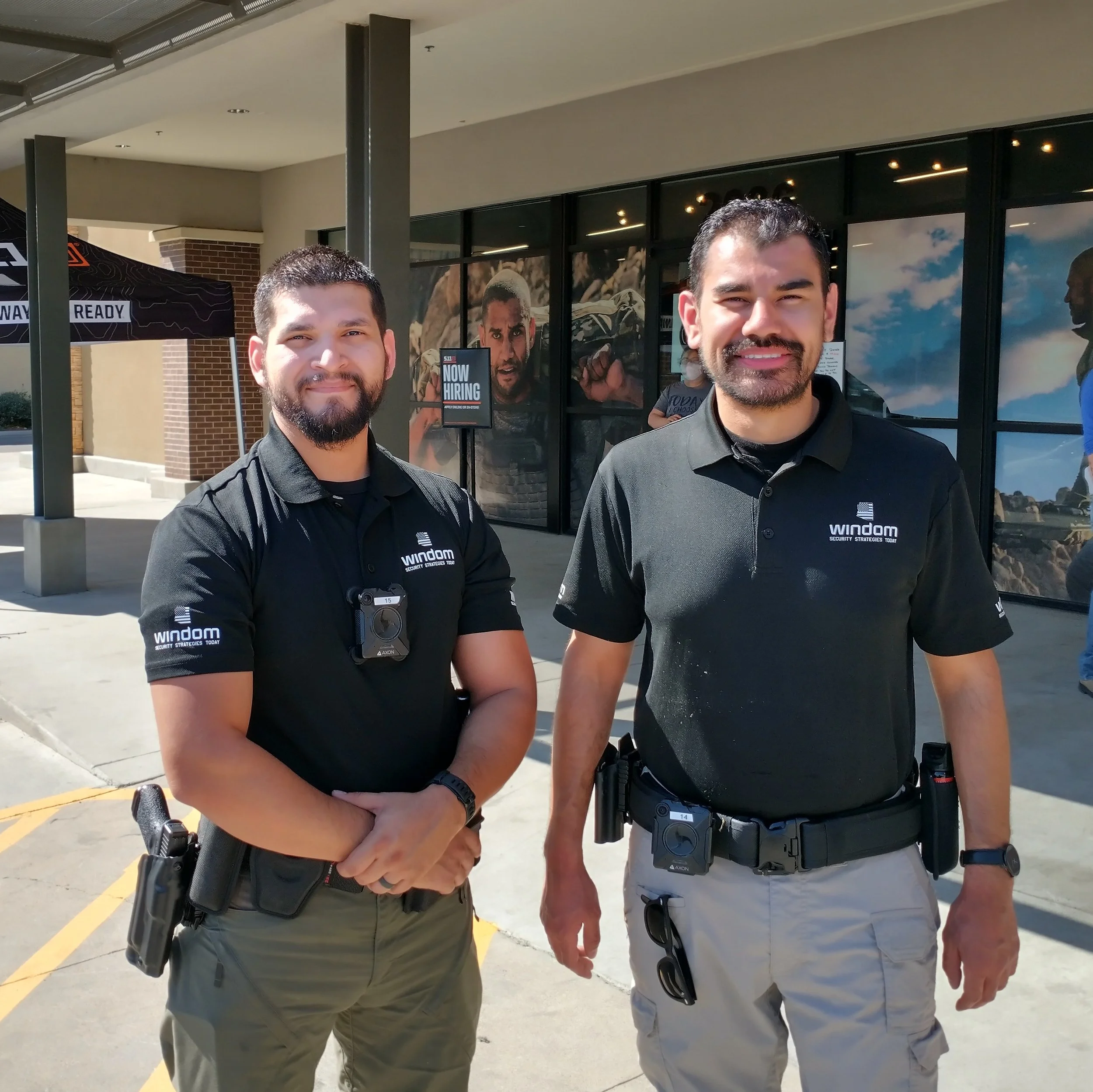 Two security guards standing outside a building, both wearing black shirts with the brand 'Windom' and security equipment, holding hands in a handshake or close gesture.