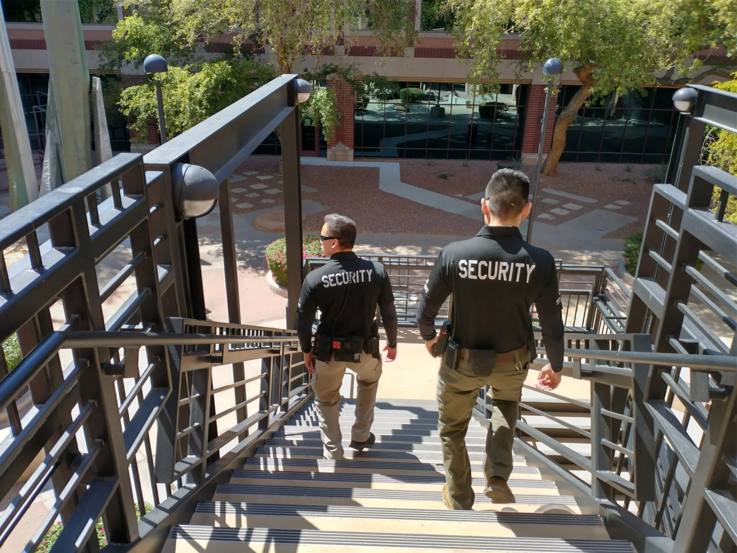Two security personnel walking down outdoor metal stairs of a building with trees and glass windows in the background.